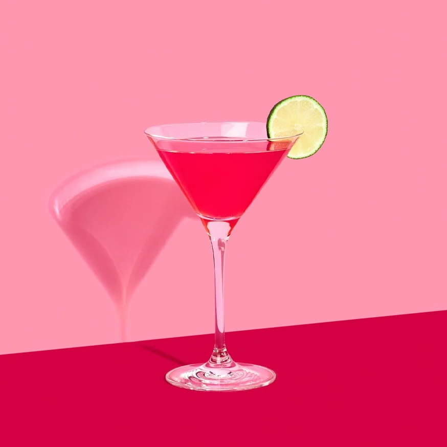Cosmopolitan cocktail med lime i martiniglas mot rosa bakgrund.
