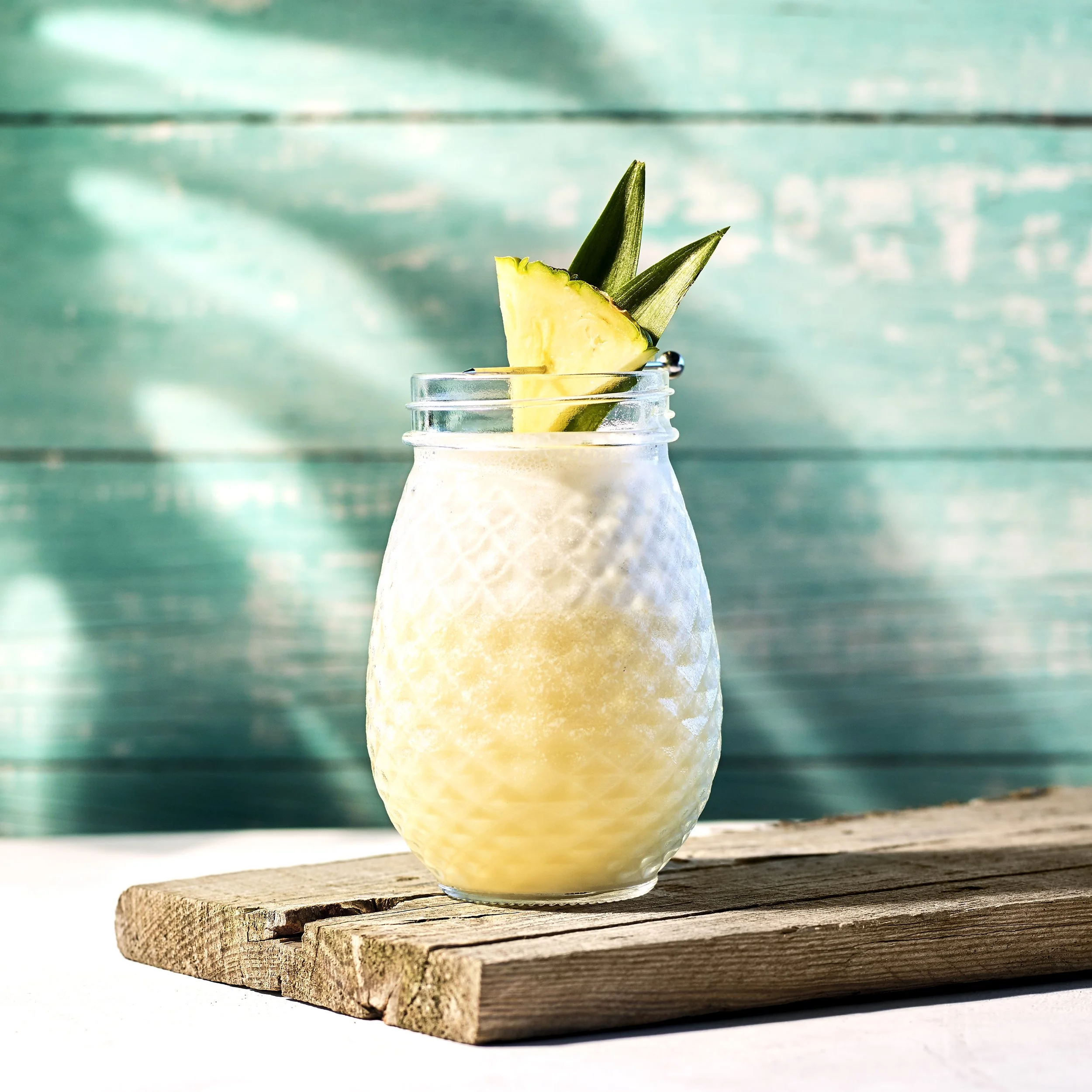 Piña Colada cocktail med ananas i tiki-glas mot turkos träbakgrund.