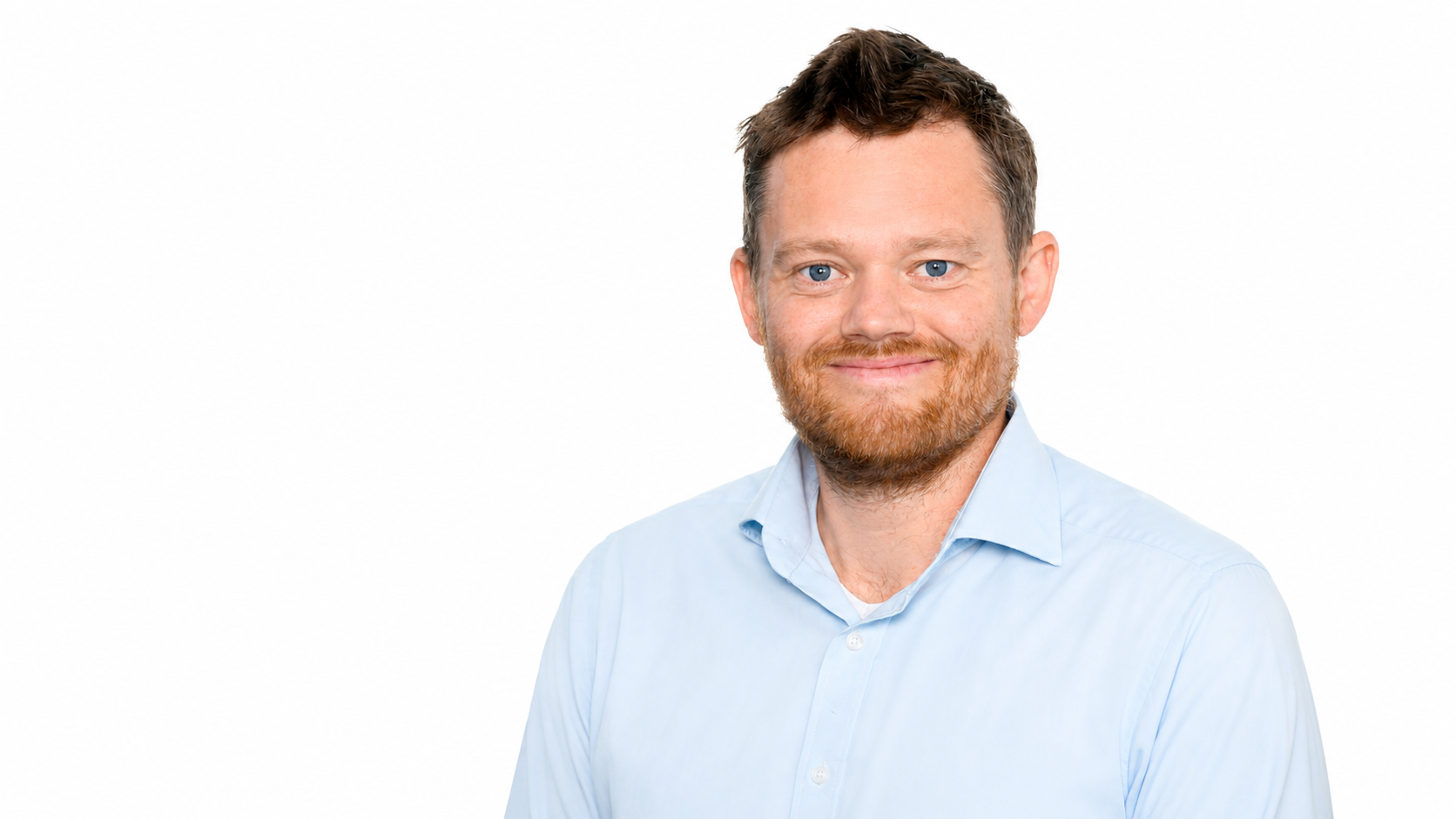 Portæt af en mand med lyseblå skjorte, kastanjebrunt hår og fuldskæg, smilende mod kameraet mod en hvid baggrund.