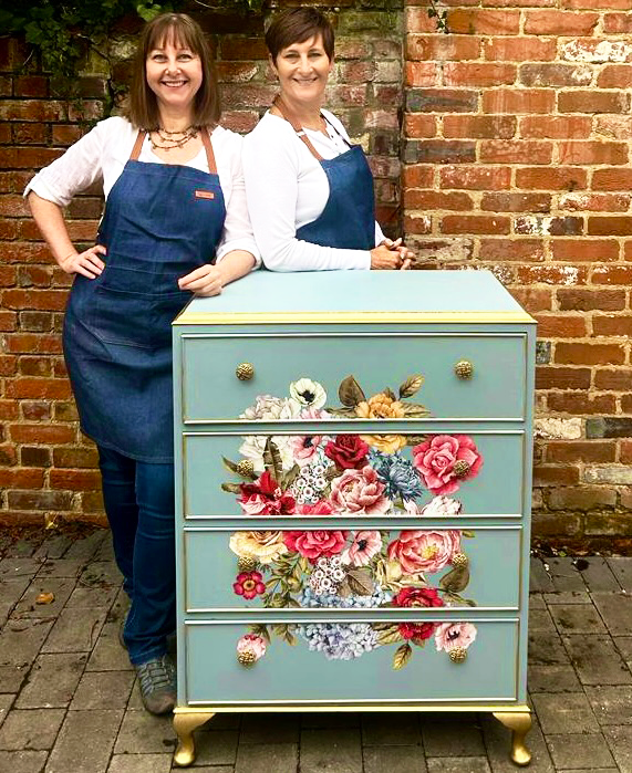 Two female furniture artists  wearing aprons standing next to a hand painted blue chest of drawers with floral decorations, positioned in front of a brick wall.
