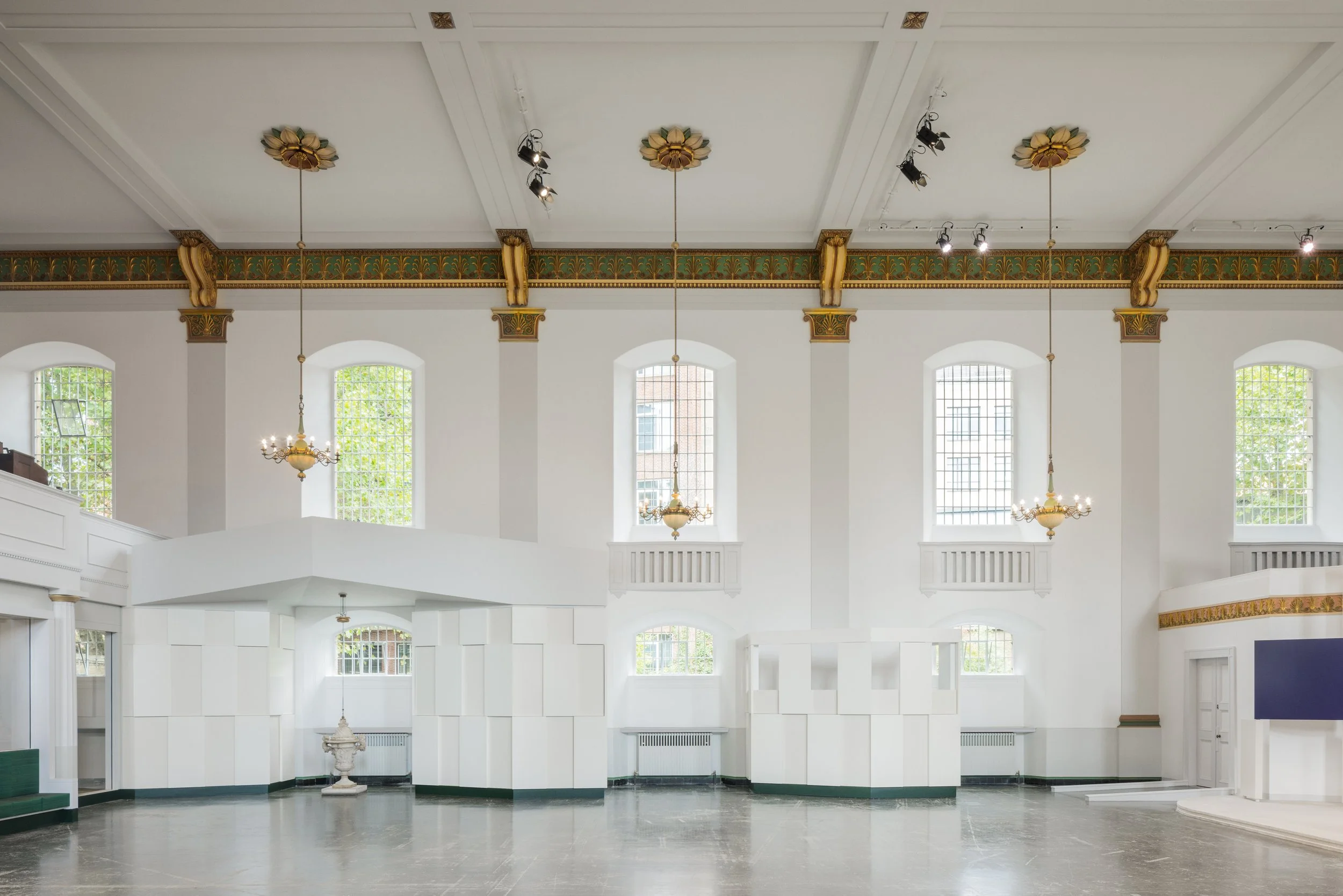Empty interior space with high ceiling, large windows with bars, white walls, decorative gold accents, chandeliers, and stage area.