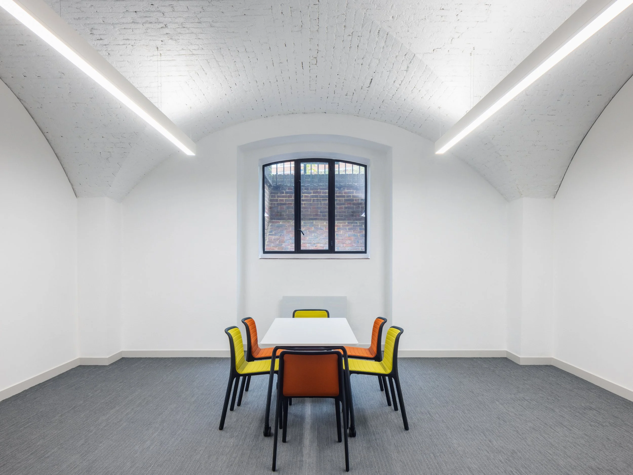 A plain white room with a curved ceiling and a gray carpeted floor. There is a window with a view of a brick wall, and a white table with six colorful chairs around it, including orange, yellow, and black chairs.