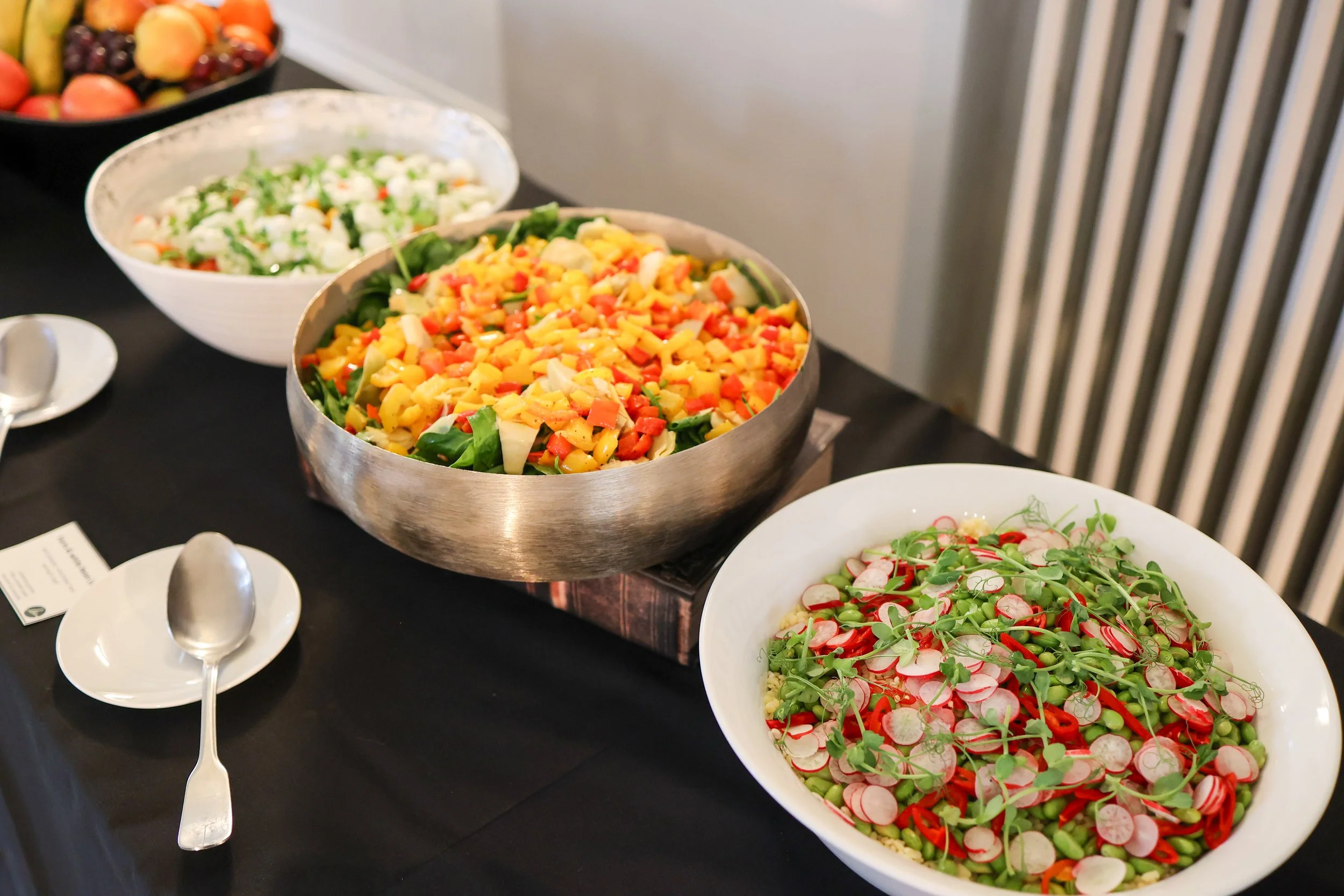 A table with three bowls of colorful salads, including a vegetable salad with diced orange, yellow, and red vegetables, a radish and green microgreens salad, and a cauliflower salad, with serving spoons and small plates nearby.