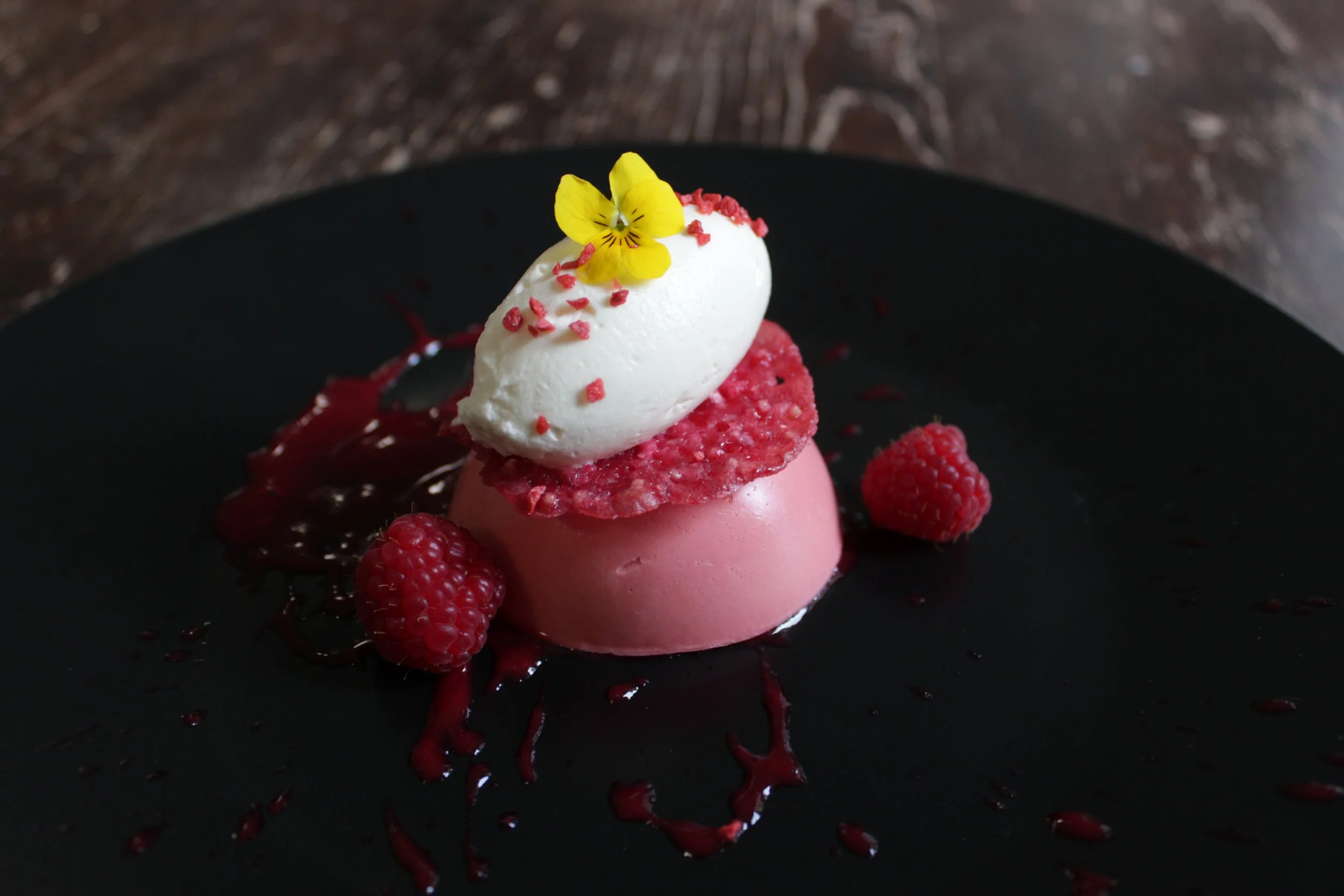 A pink and white plated dessert with raspberries, raspberry sauce, and a yellow edible flower.