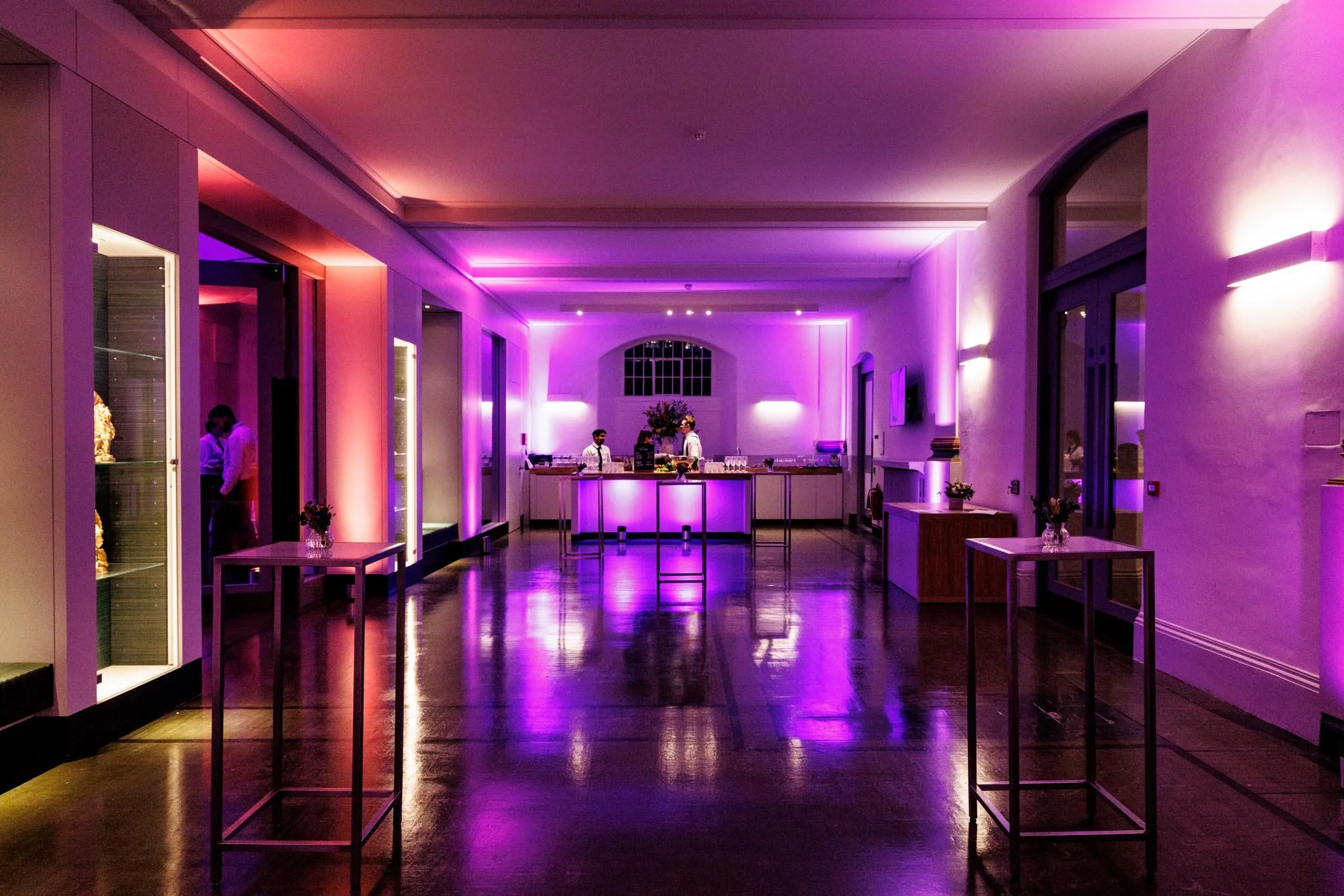 Interior of a dimly lit event space with pink and purple lighting, tables with flower arrangements, a bar area at the back, and a couple of people in conversation on the left side.