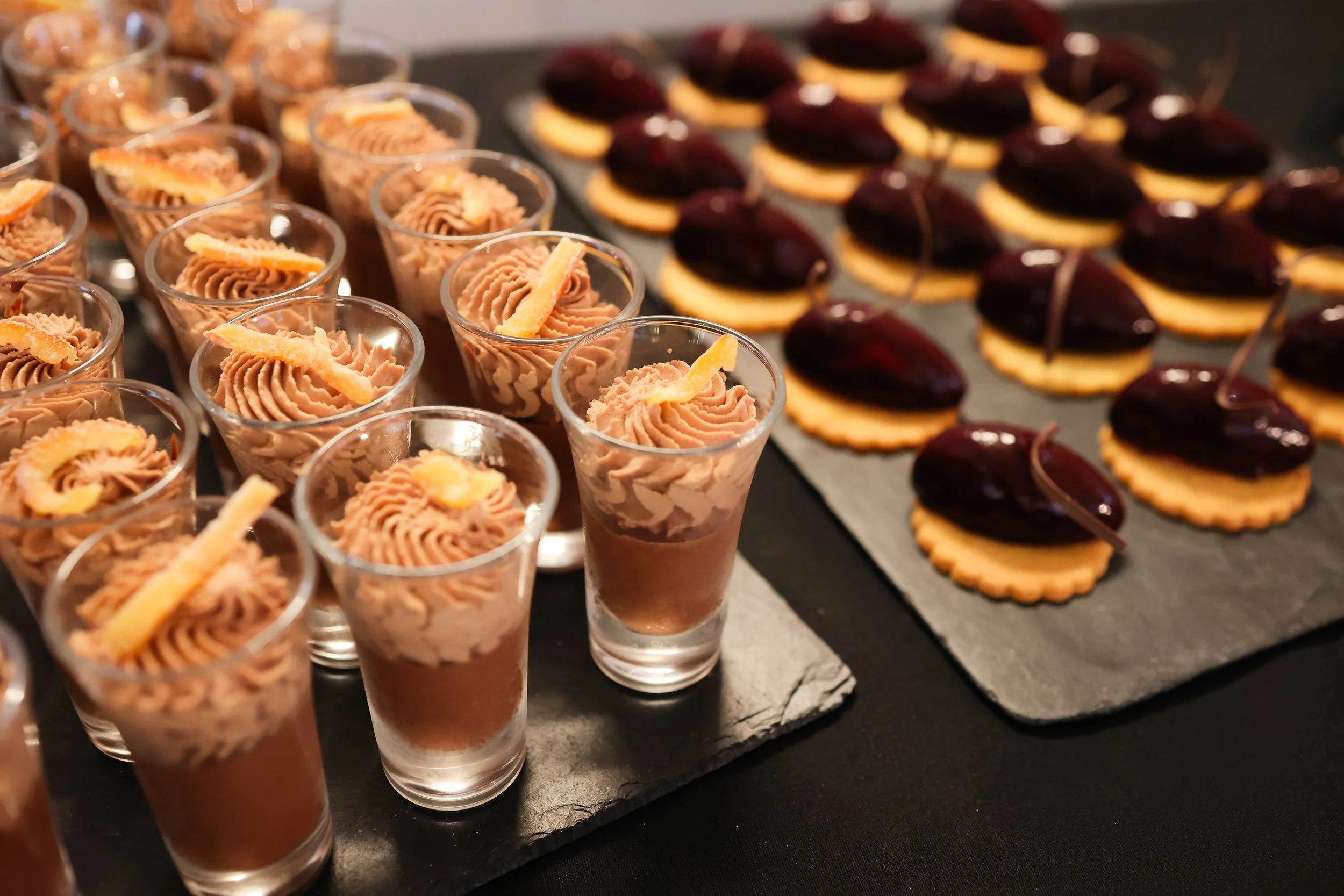 Assorted desserts on black slate serving trays, including mousse in small glasses topped with garnishes and finger foods with cherries and cookies.