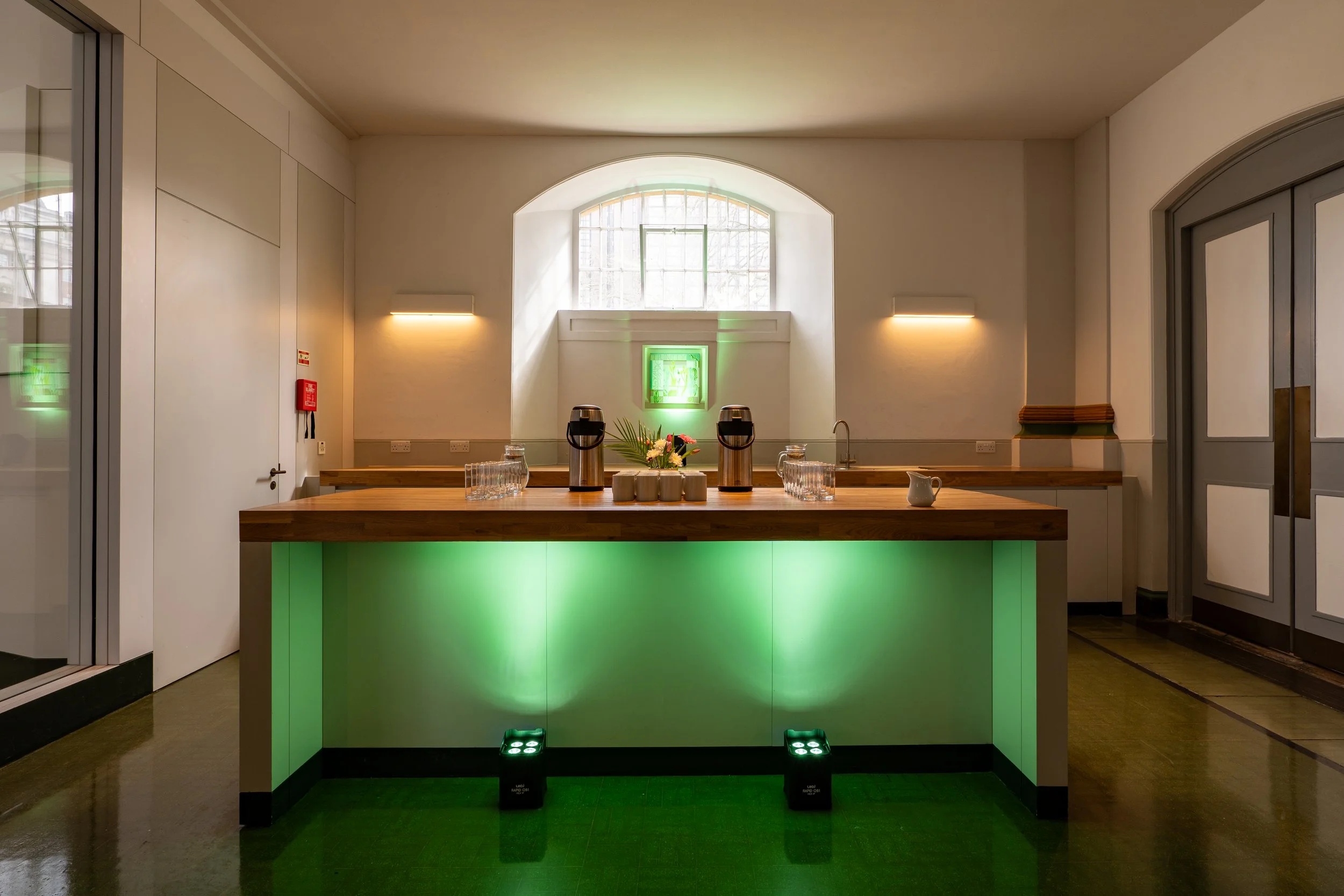 A modern indoor space with a green-lit bar counter, two coffee dispensers, glasses, and a small pitcher on top, illuminated by wall-mounted lights and natural light from a window.