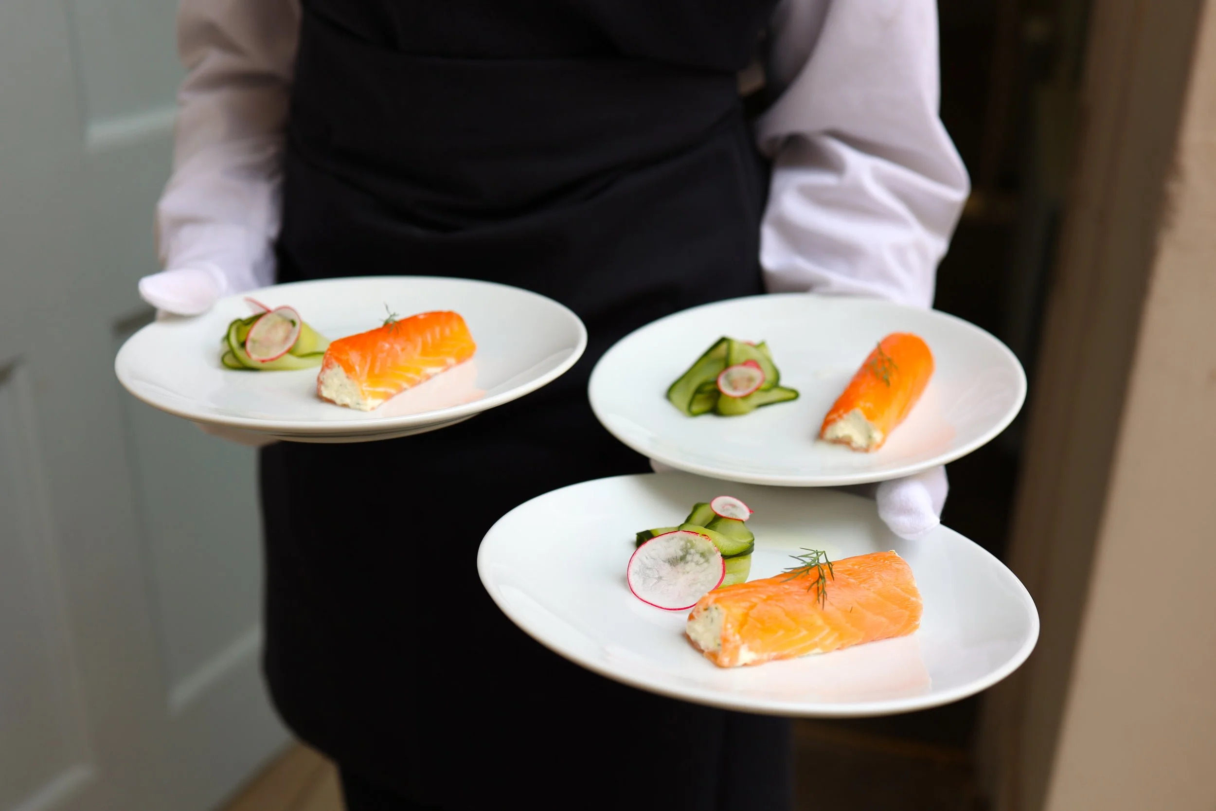 A waiter holding three white plates with smoked salmon, cucumber slices, radish slices, and dill garnishes.