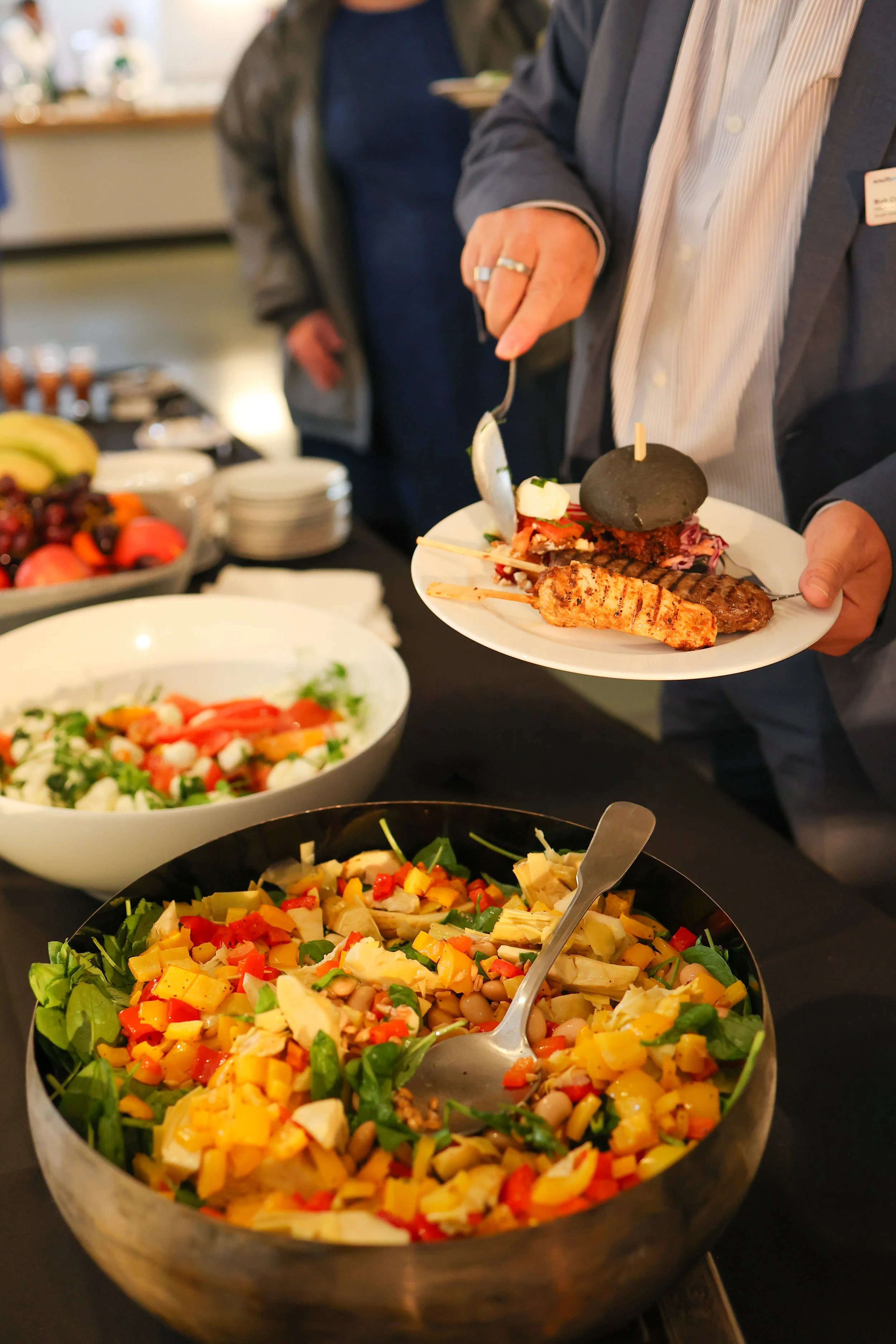 A person holding a white plate with grilled chicken, a burger with a black bun, and grilled meat skewers, at a food buffet with salads and fruit.