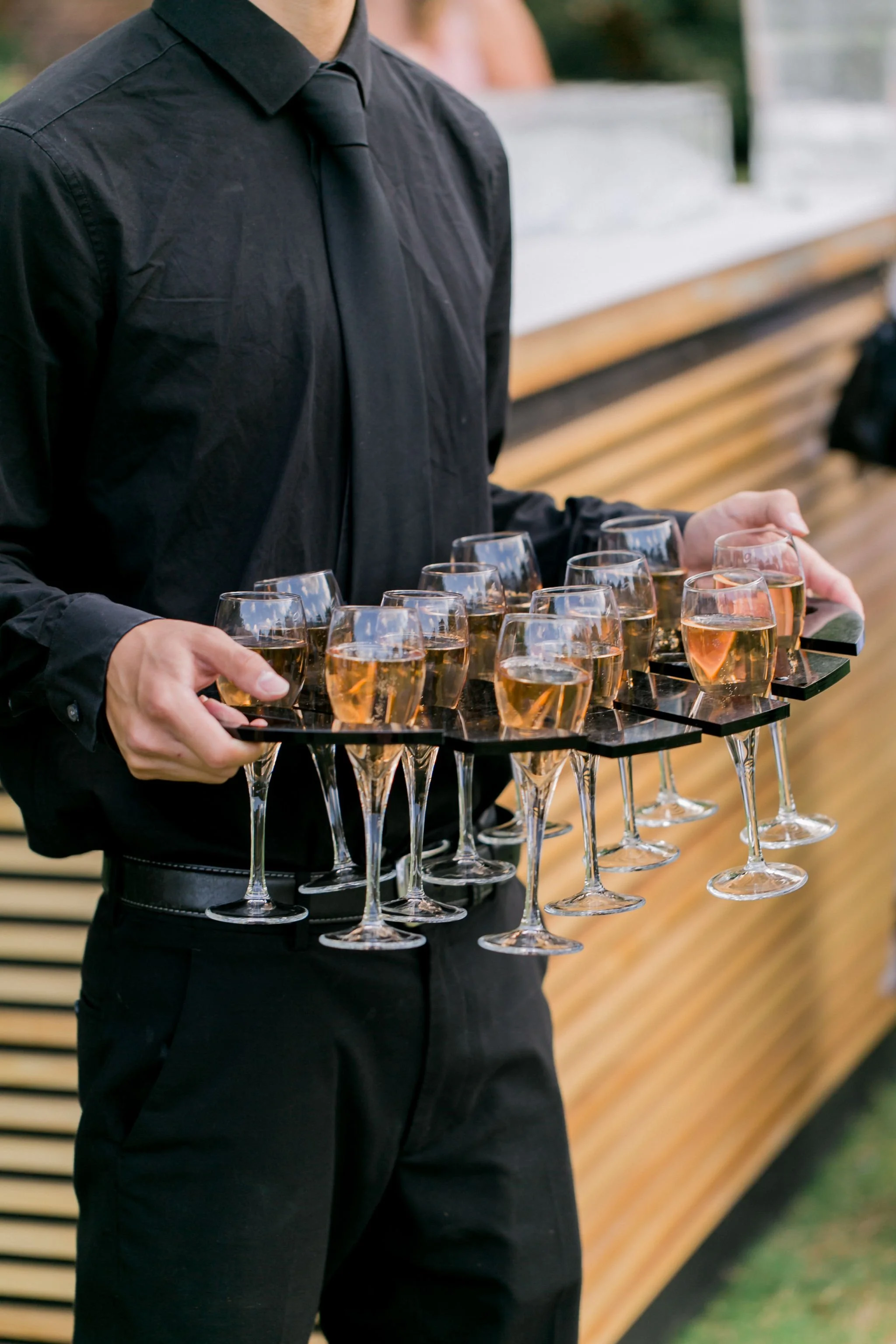 Person dressed in black holding a tray of champagne flutes filled with rosé champagne at an outdoor event.