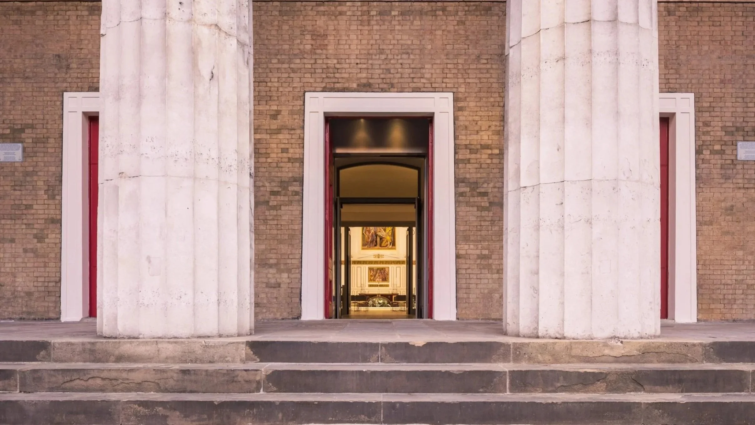 Exterior view of a classical building with brick walls, large white columns, and steps leading to a doorway. Through the open door, an interior with paintings and a room is visible.