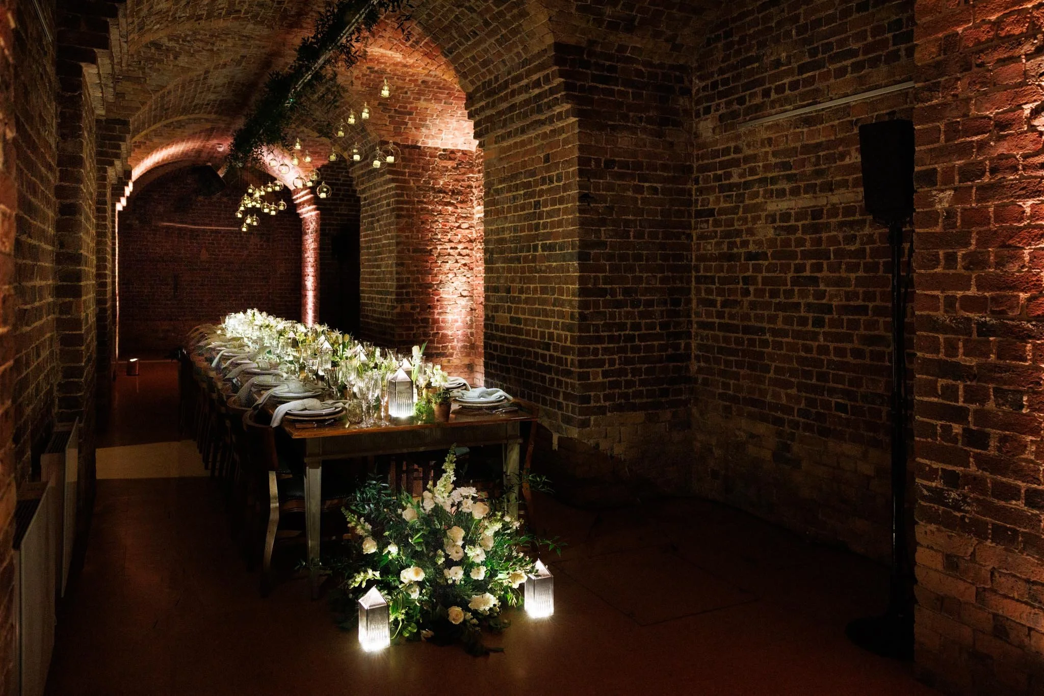 A long dining table set for a formal event with white flowers and candles, decorated with hanging lights and greenery, against brick walls and arched ceilings.