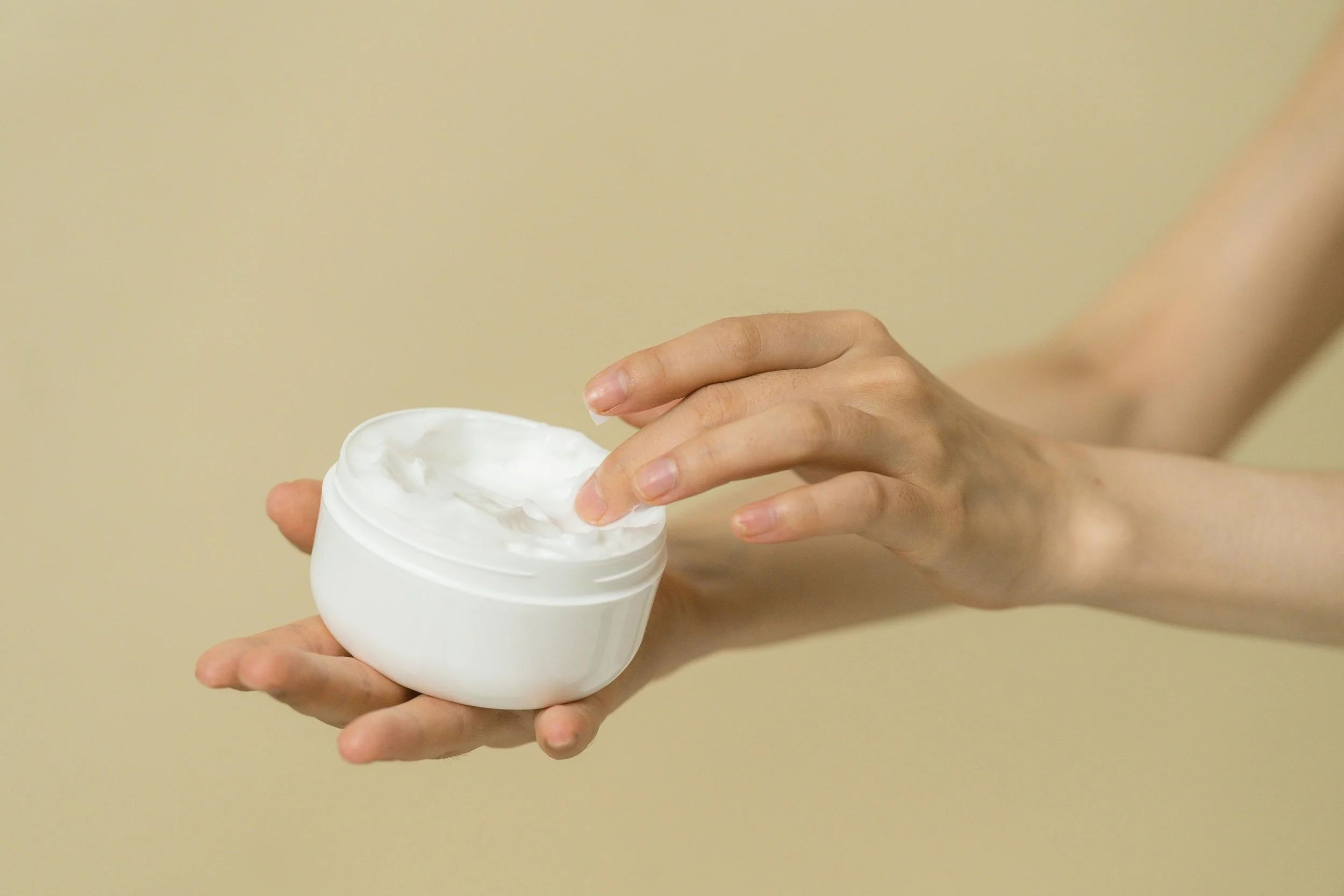 Hand applying a rich moisturizing cream from a white jar on a soft beige background, highlighting skincare and beauty routine.