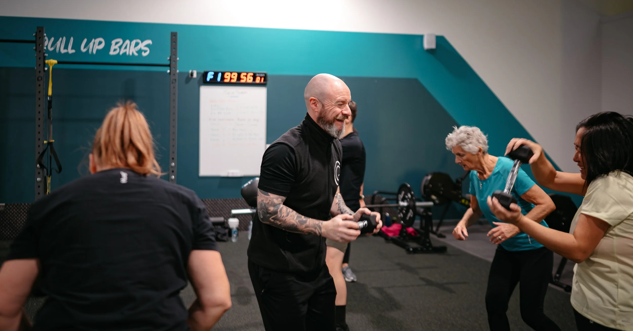 A group of diverse older adults and a trainer enjoy a lively workout class in a gym, with weights and exercise equipment visible in the background.