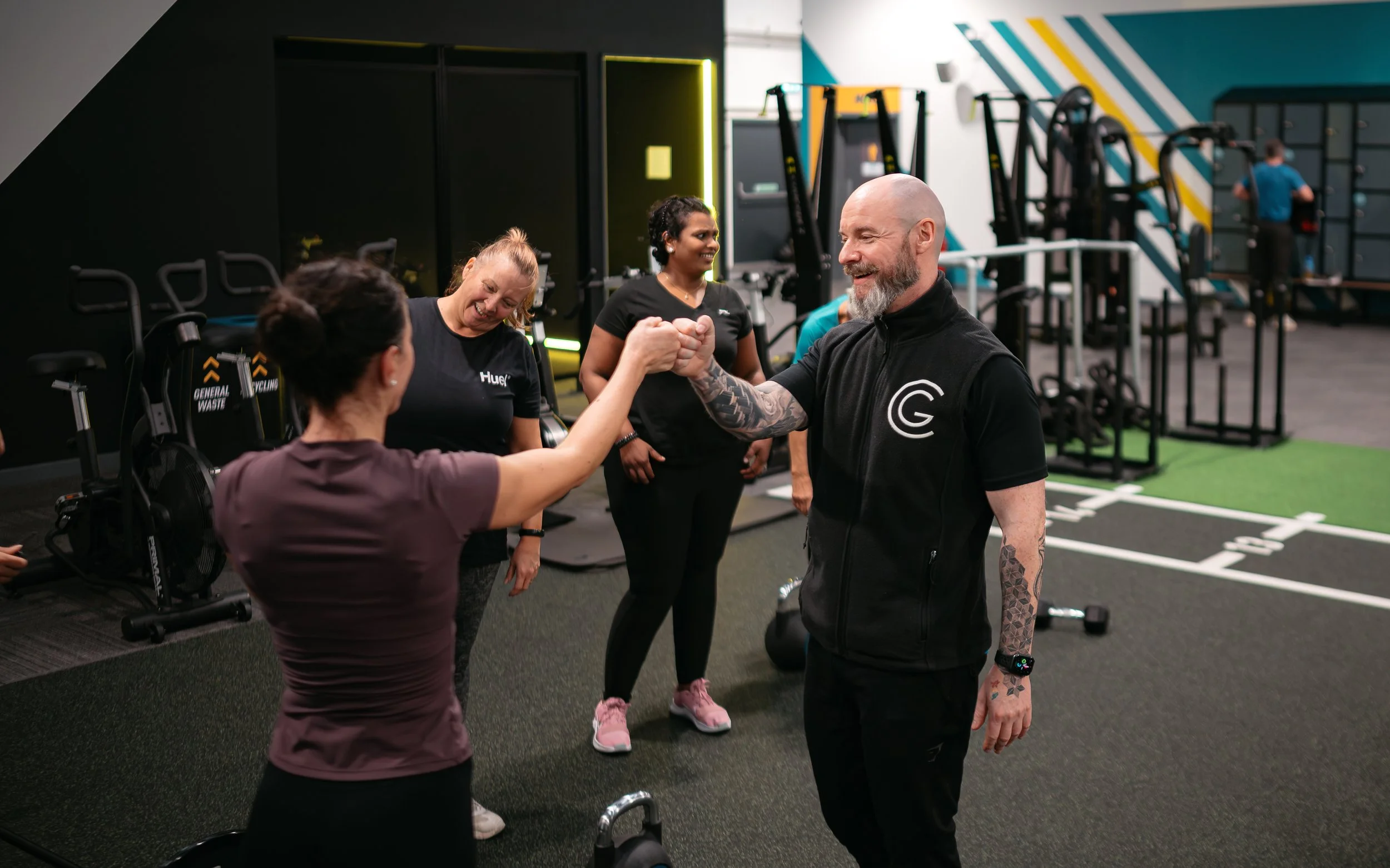 Two people, a woman and a man, are greeting each other with a fist bump in a gym. Other gym members are visible in the background, along with workout equipment and a track area.