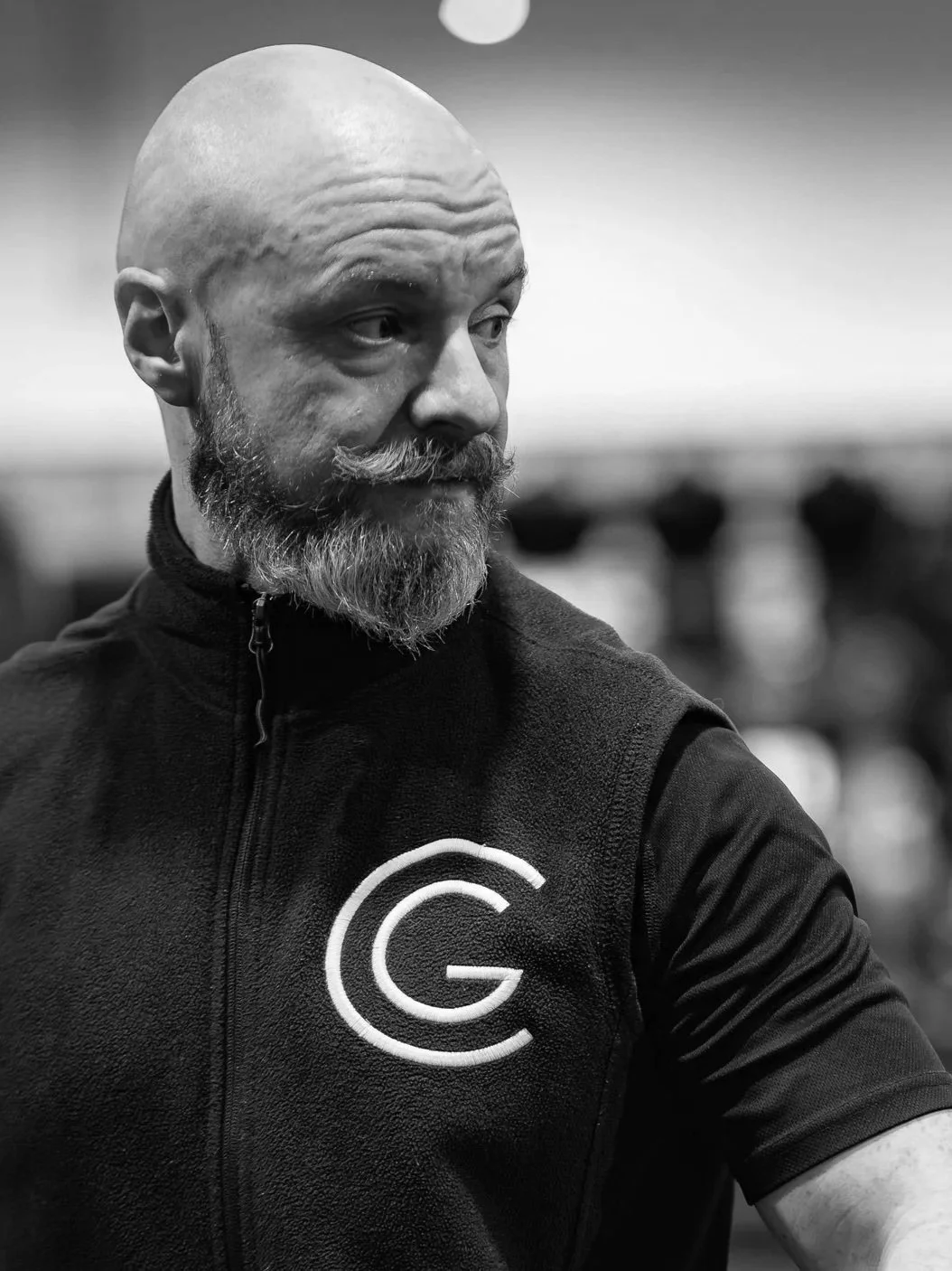 A black and white photo of a man with a bald head and a beard, wearing a black athletic jacket with a logo on the chest, looking to his right.