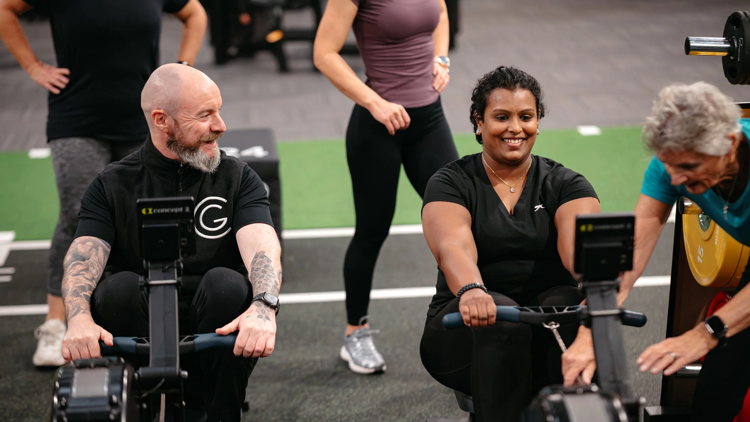 People exercising on rowing machines at a gym, smiling and engaging with each other.
