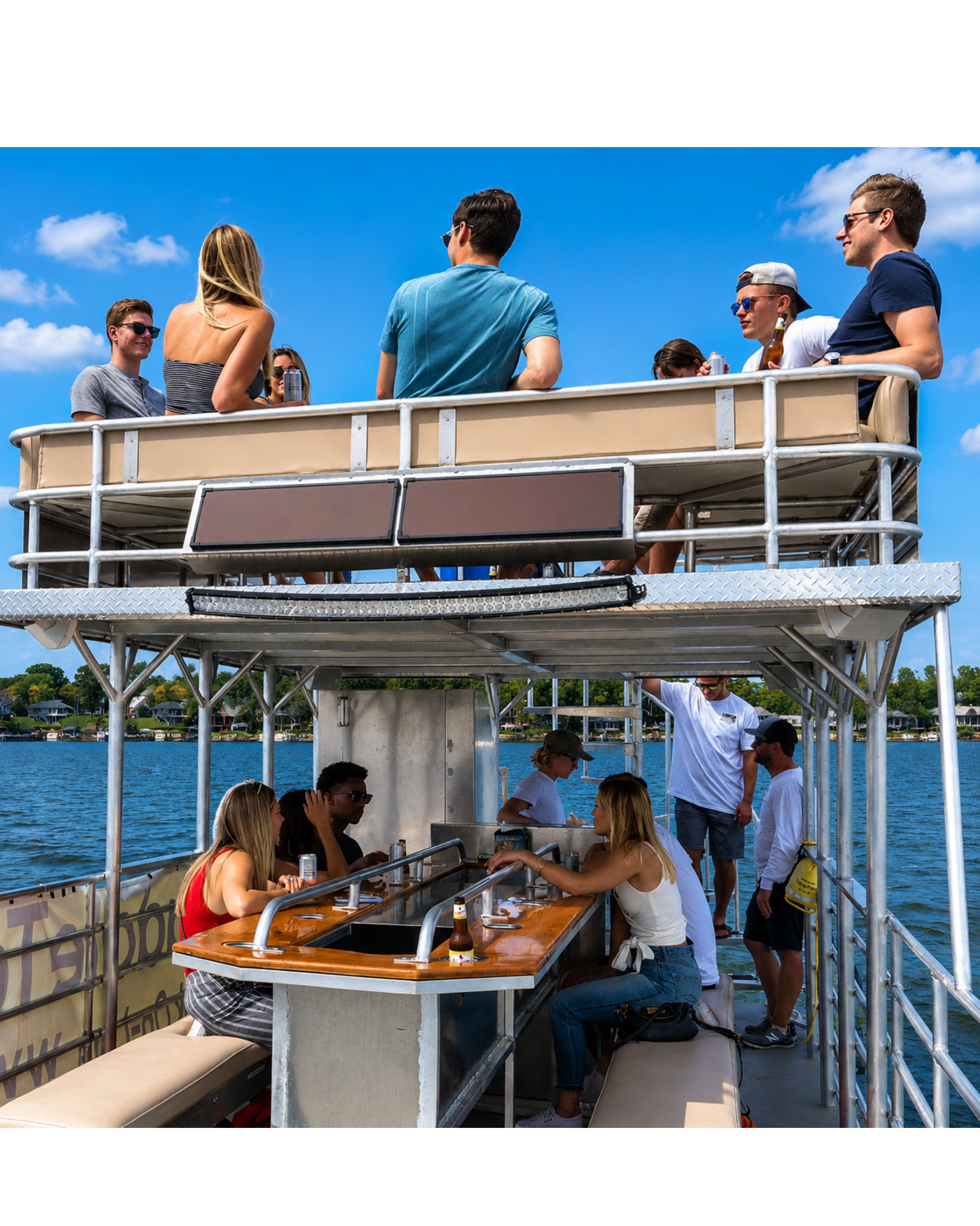 People enjoying a boat ride on a sunny day, sitting around a table with drinks, some standing, with a view of water and trees in the background.