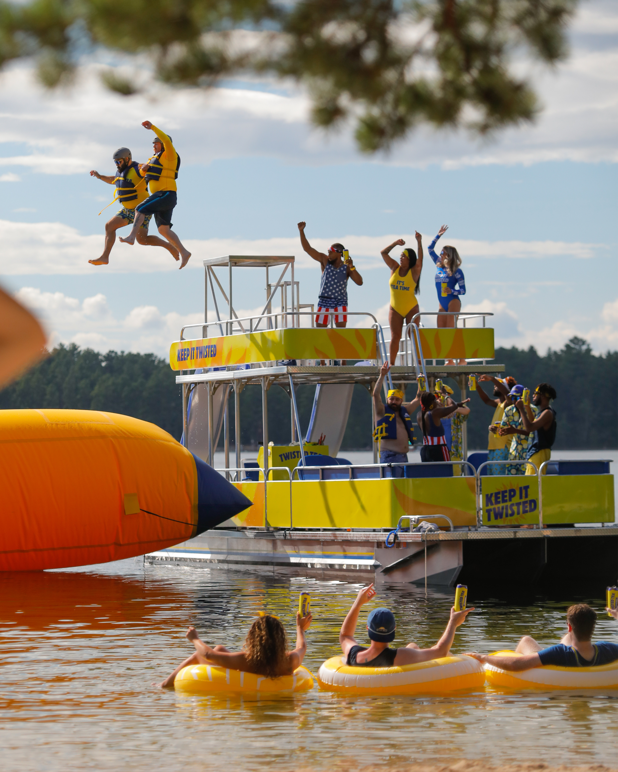 People in life jackets jumping from a floating dock into a lake during a summer event, with spectators in the water holding signs.
