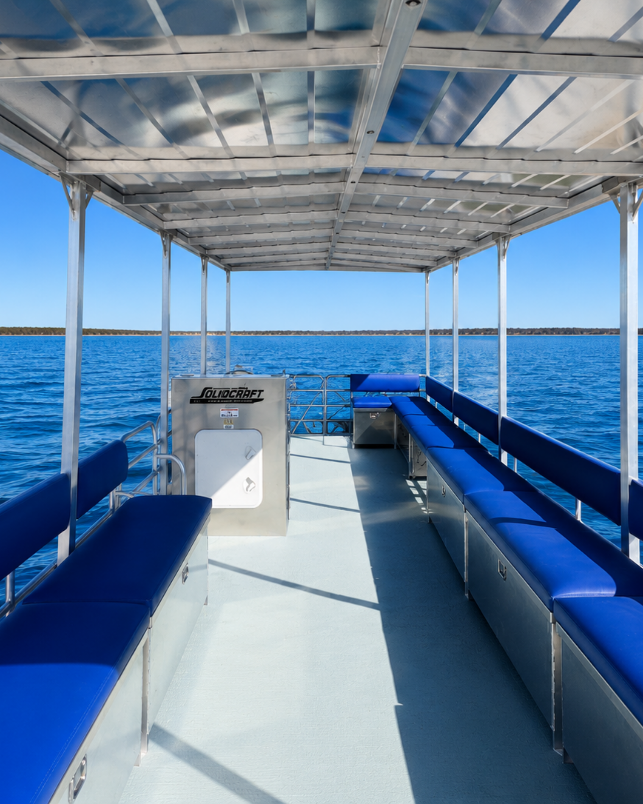 View from the deck of a boat with blue cushioned seating along the sides, looking out over a body of water with a landmass in the distance under a clear blue sky.