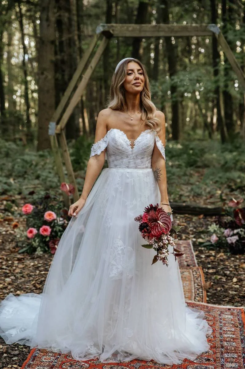 Bride with soft waved hair  and soft glam makeup stands in woodland holding a bouquet