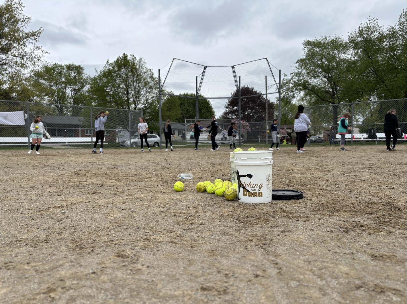 A group of people practicing softball on a dirt field, with baseballs and a bucket labeled 'Pitching with Dana' in the foreground, and trees and a cloudy sky in the background.