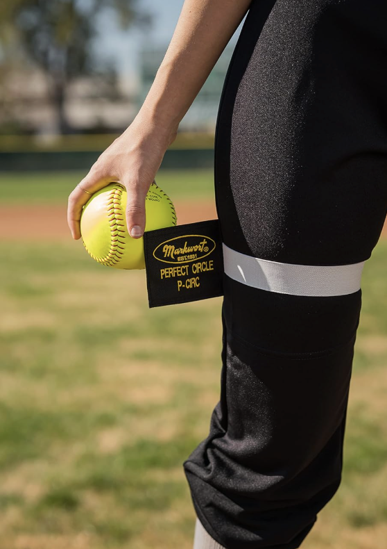 Close-up of a person holding a yellow softball in their left hand, standing on a baseball field, wearing black athletic pants with a white stripe, and a black strap labeled 'Markwort Perfect Circle P-CIRC' around their thigh.