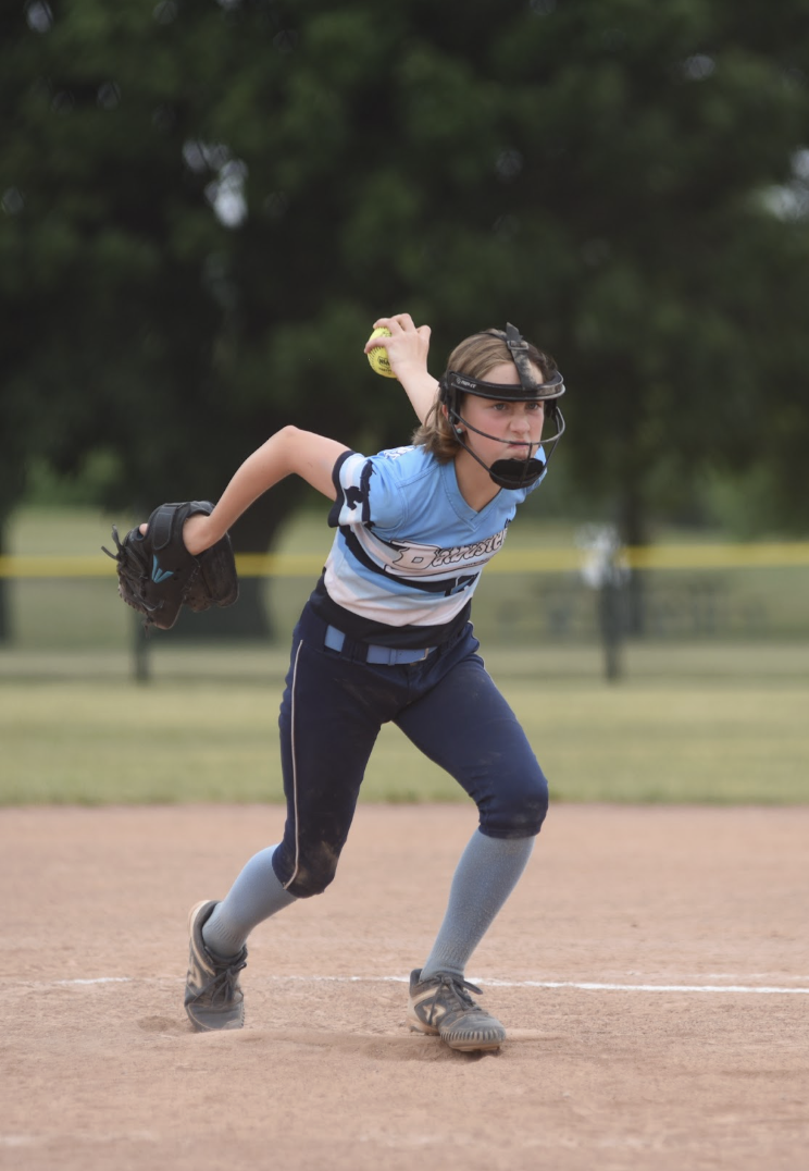 A young girl playing baseball on a dirt field, wearing a blue uniform, gray socks, and a black helmet with a face guard, in a crouched position preparing to throw a yellow baseball.