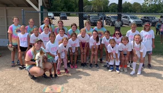 Group of young girls in sports uniforms and t-shirts on a baseball field, some holding a dog, with a chain-link fence and parked cars in the background.