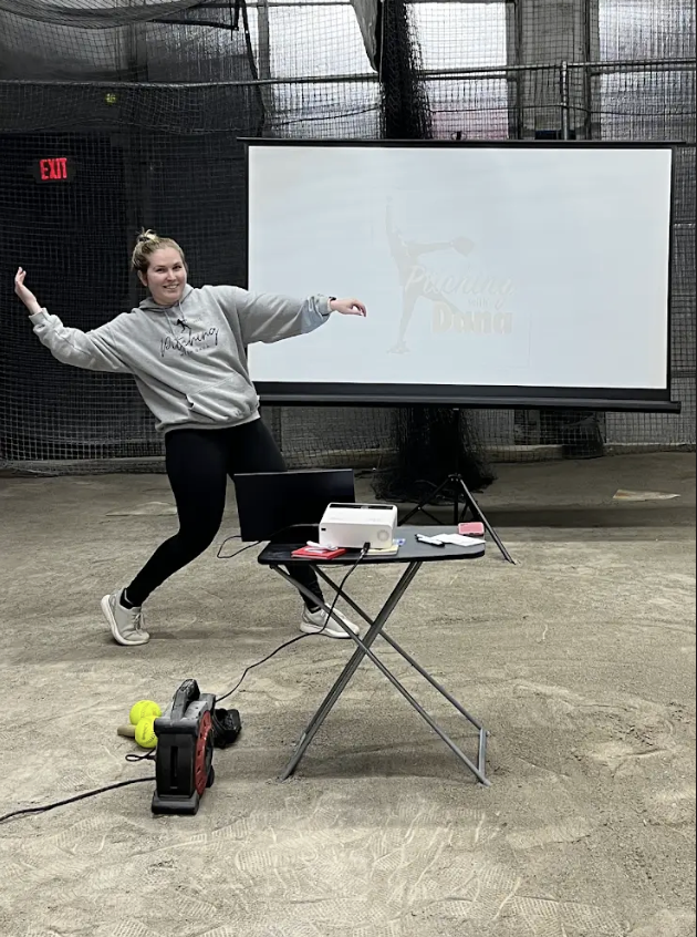 A woman in athletic clothing stands in front of a large screen, striking a playful pose with arms extended. She is indoors, in what appears to be a sports or training facility, with gym equipment and tennis balls on the floor nearby. A table with a laptop and projector is set up in front of her.