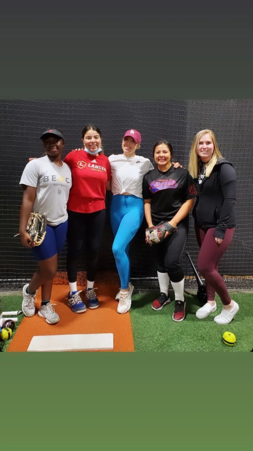 Group of five young women standing together inside a softball training facility.