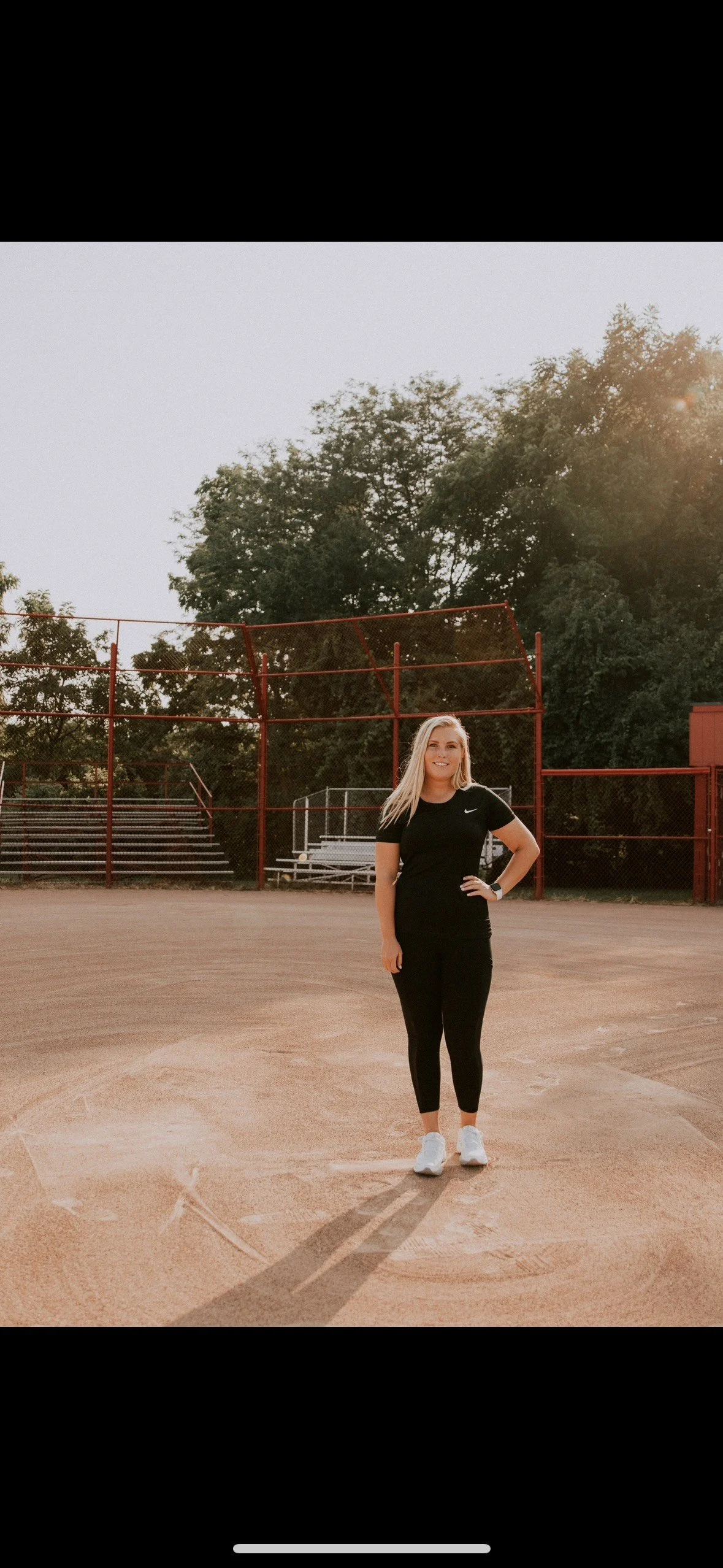 A woman in black athletic clothing standing on a baseball field with trees and empty bleachers in the background.