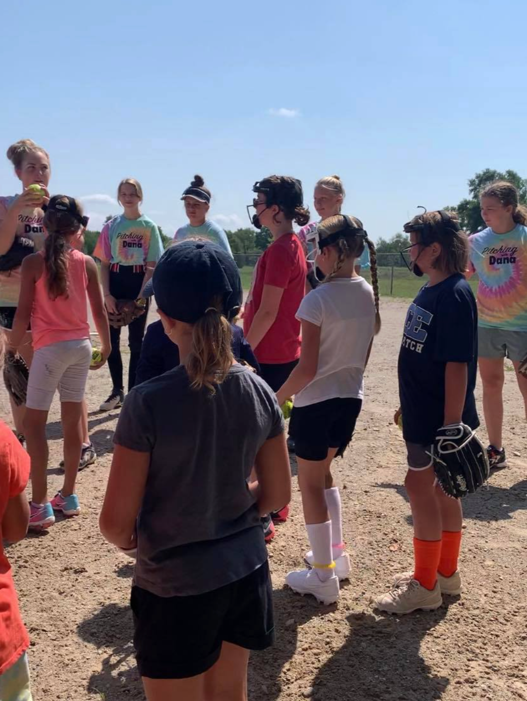 A group of young girls gathered outdoors on a sunny day, some wearing sports uniforms and holding softball gloves and balls, appear to be participating in a softball practice or game.