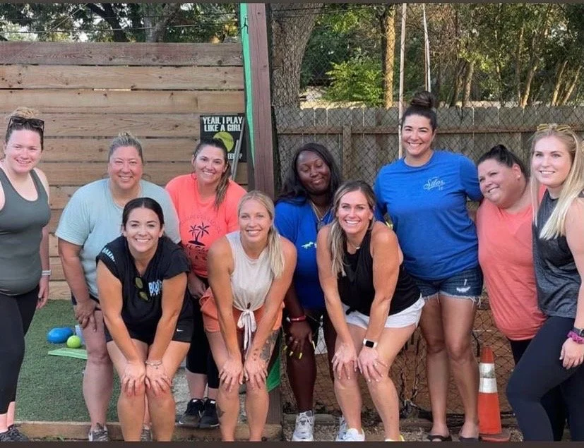 Group of nine women posing outdoors during daytime, smiling, with a wooden fence, trees, and a sign in the background.