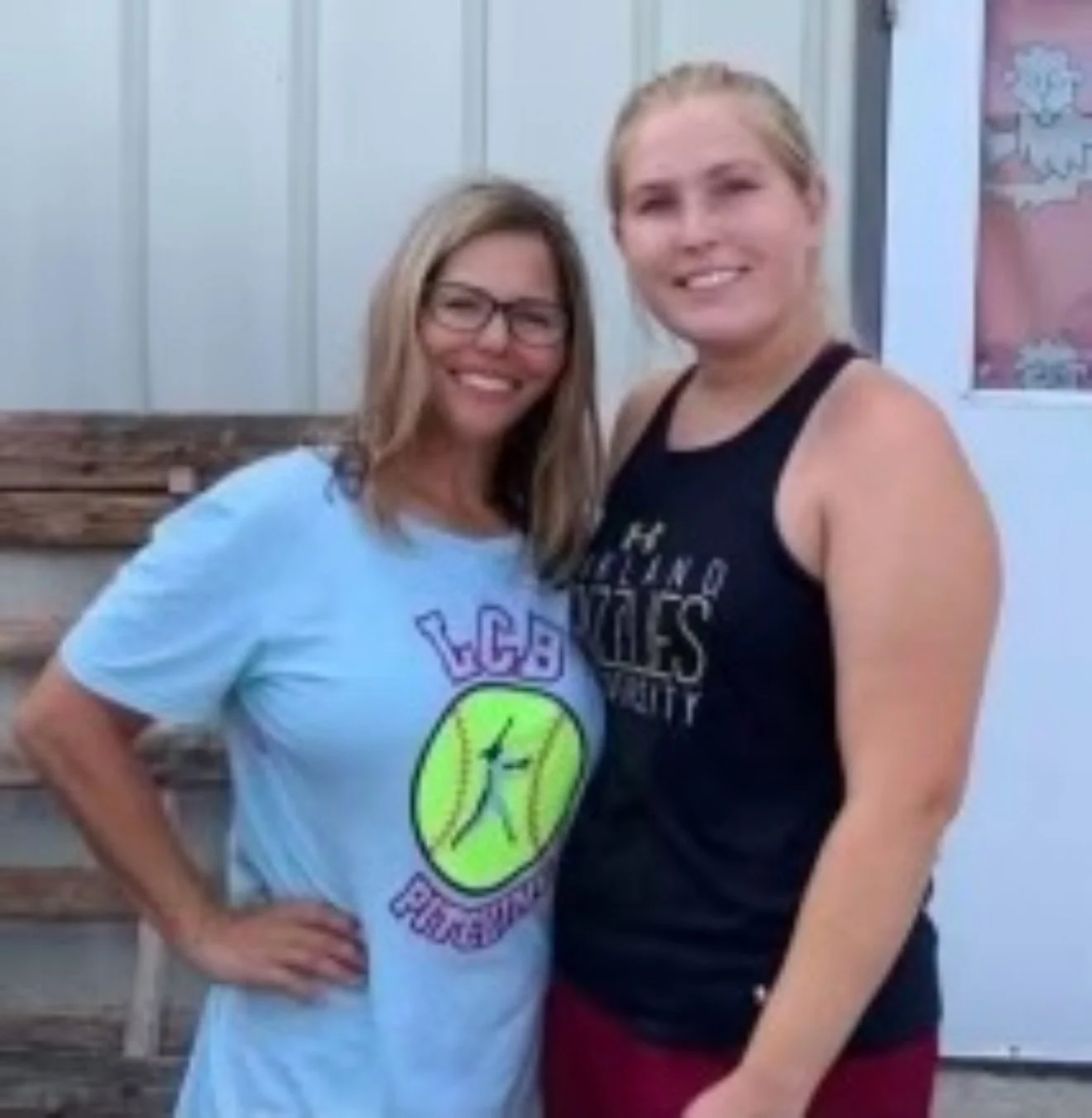 Two women standing outdoors, smiling and posing for a photo, with a wooden fence and a sign in the background.