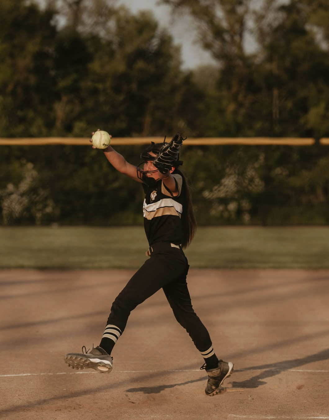 A female softball player is in mid-air on a softball field, holding a ball in her right hand and wearing a catcher's mask pushed up onto her head, with a glove on her left hand. She appears to be celebrating or preparing to throw the ball.