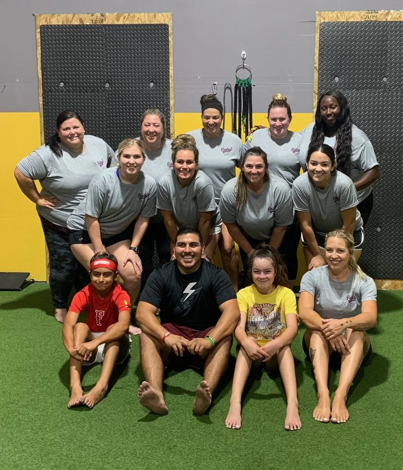 Group of nine women and three children in a gym, with some on the floor and some standing, all smiling. The background includes gym equipment and black padded walls.