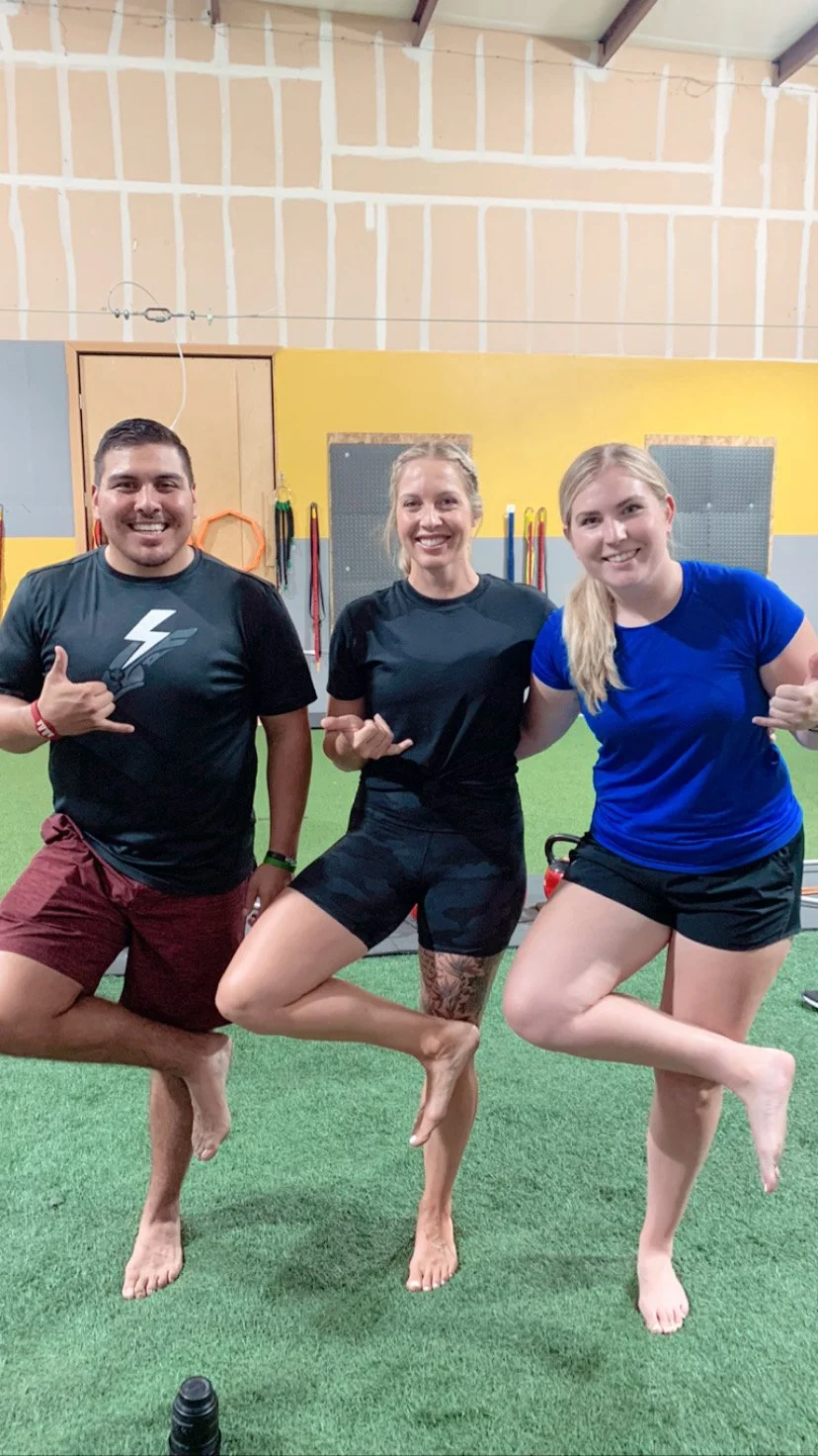 Three people smiling and making a shaka sign with their right hands, standing on one leg with the other leg bent and raised, inside a gym with green turf flooring and gym equipment in the background.