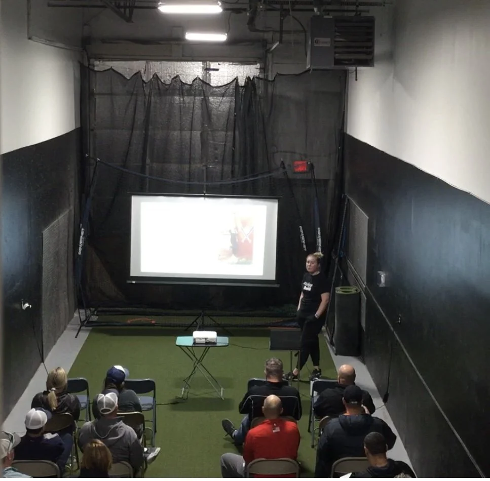Indoor presentation with a woman speaking to a seated audience, a projector screen displaying a slide, and black protective netting on the walls.