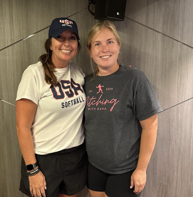 Two women smiling, one wearing a USA softball cap and t-shirt, the other in a gray t-shirt with pink text, standing side by side against a wooden wall background.
