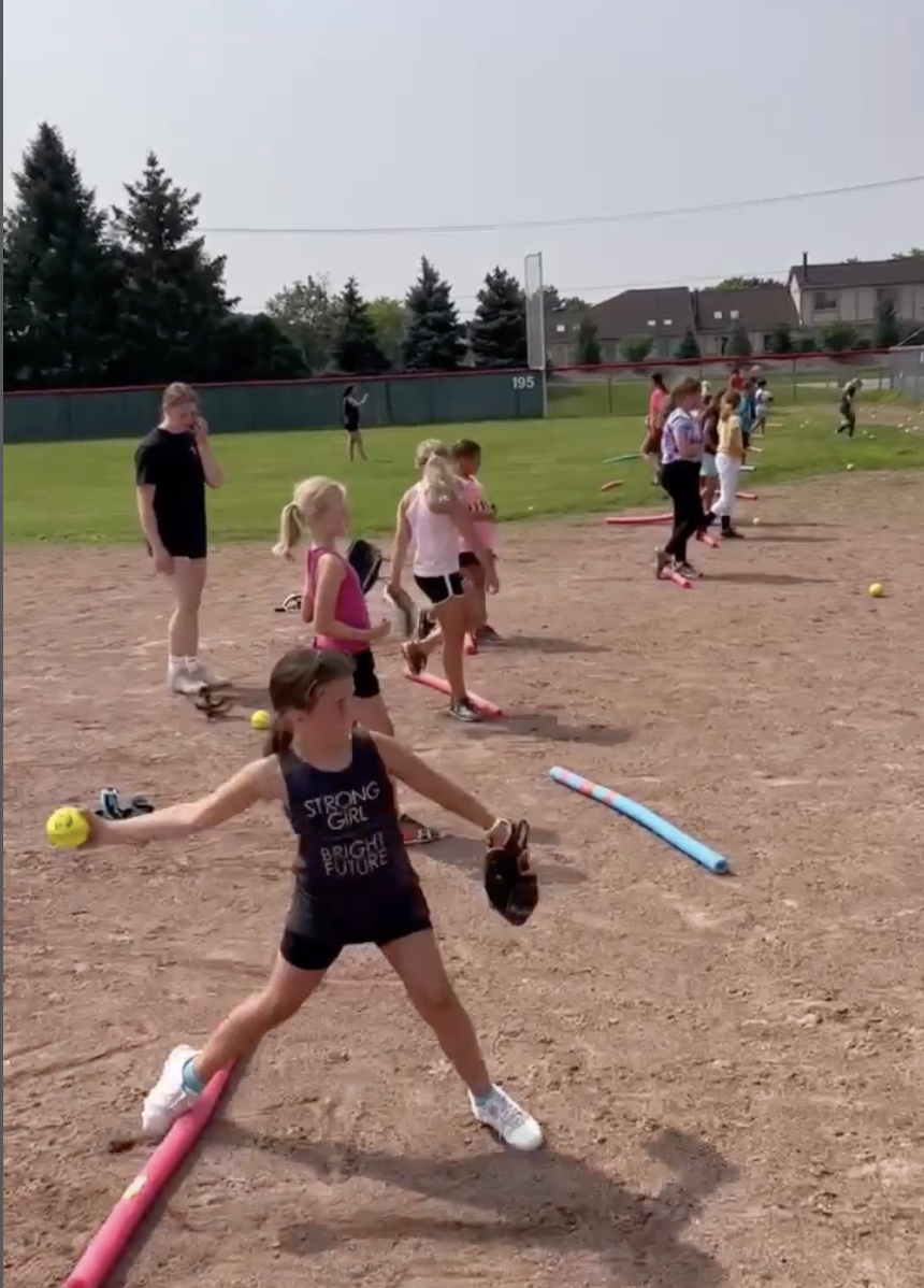 Children participating in a baseball practice on a field, with a girl in the foreground holding baseballs and a glove, and other children lined up behind her, some holding bats, on a dirt infield with a grassy outfield and trees in the background.