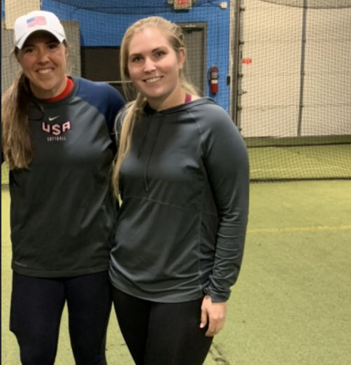 Two women standing in an indoor sports facility, smiling at the camera. One is wearing a USA softball jacket and cap, the other a dark athletic hoodie and leggings.