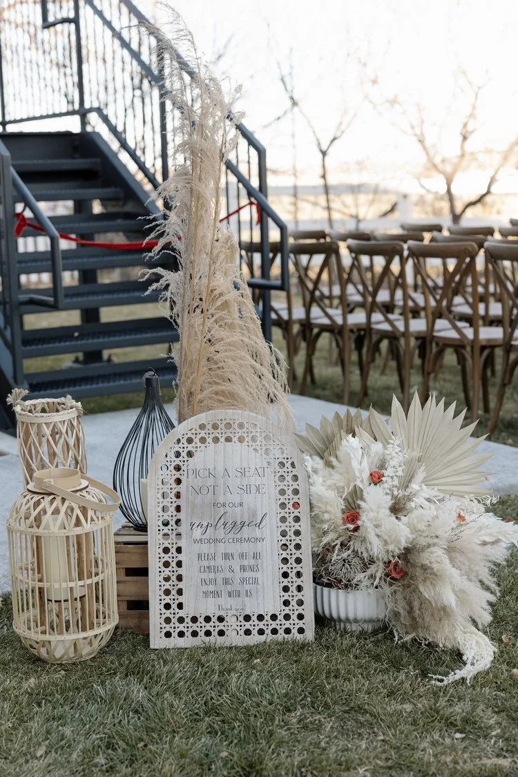 Outdoor wedding setup with decorative sign "Pick a seat, not a side" and pampas grass arrangements near seating area.