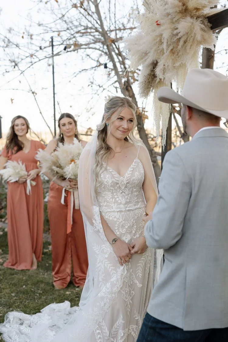 Bride in elegant gown looking at groom in cowboy hat during outdoor wedding ceremony with bridesmaids in background holding bouquets.