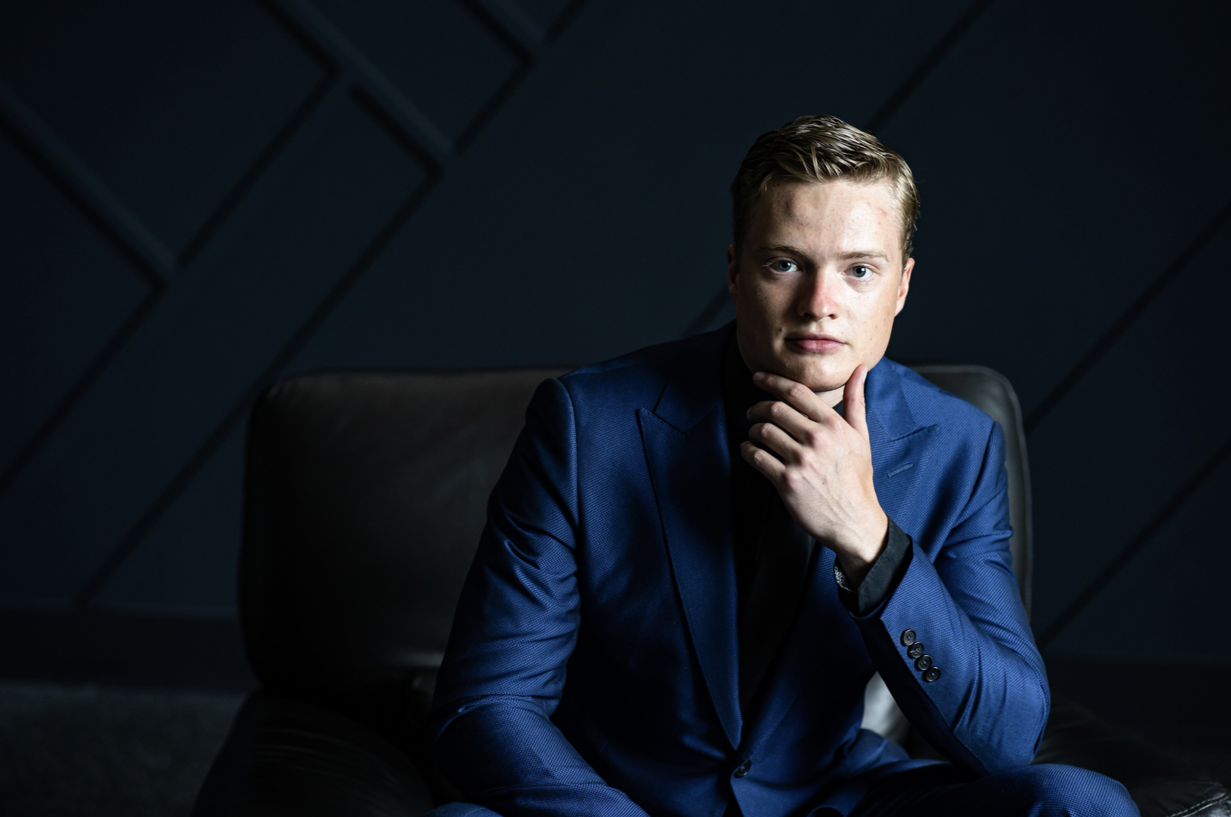 A young man with blonde hair in a blue suit sitting on a black chair, resting his chin on his hand, in a dark, modern room.