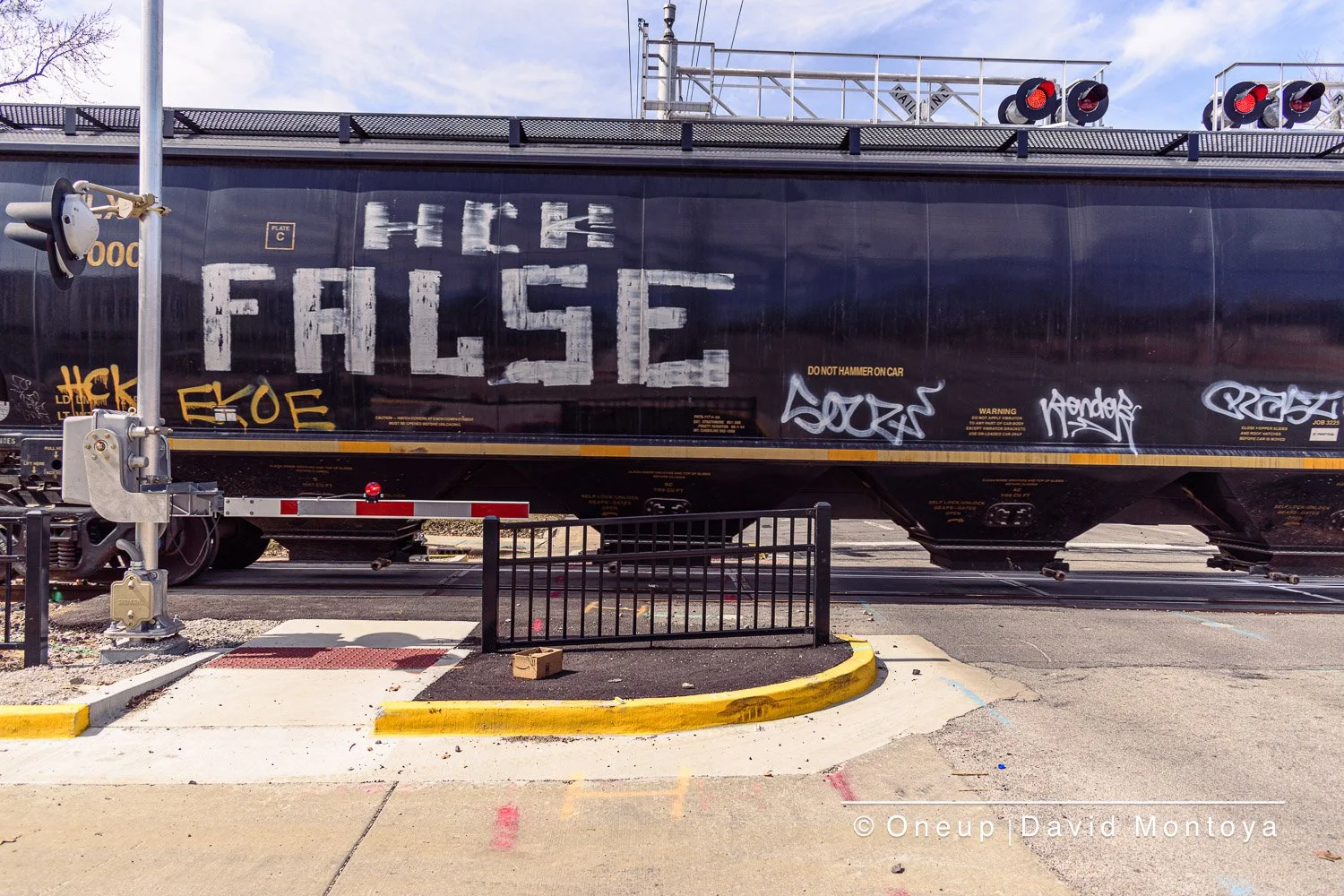 A train car with large white graffiti lettering that says 'HIGH FALISE' on the side. The train is black and sitting on tracks at a rail crossing, with a small black fence and a red-and-white barrier in the foreground. There are red lights on top of the train car. The sky is partly cloudy.
