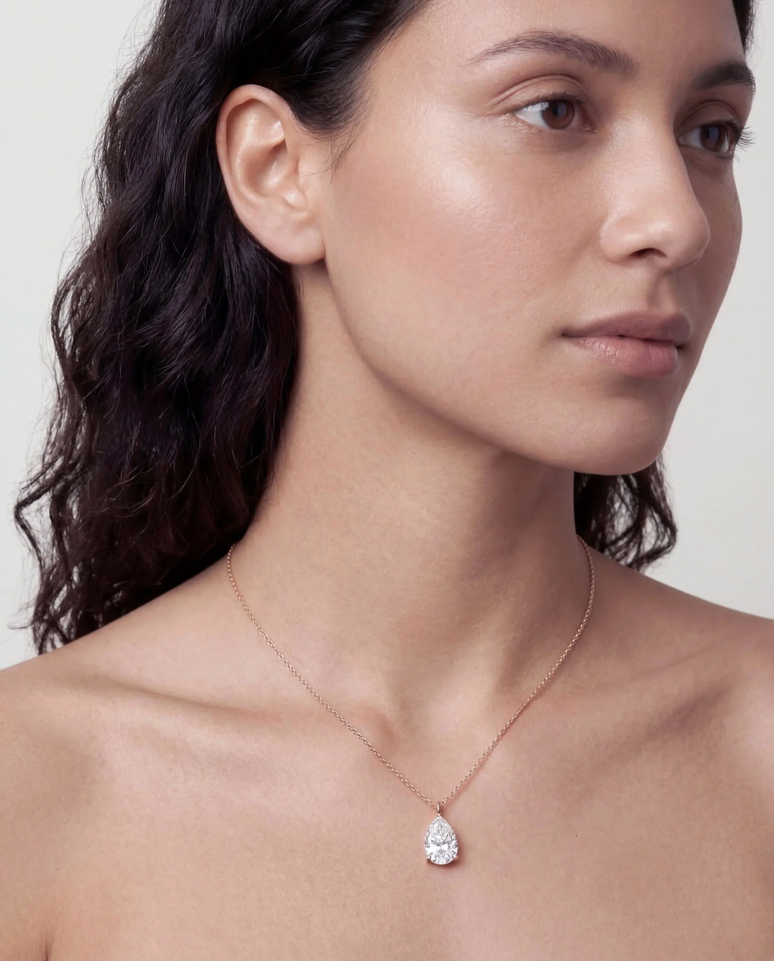 A woman with dark wavy hair wearing a delicate necklace with a teardrop-shaped diamond pendant.