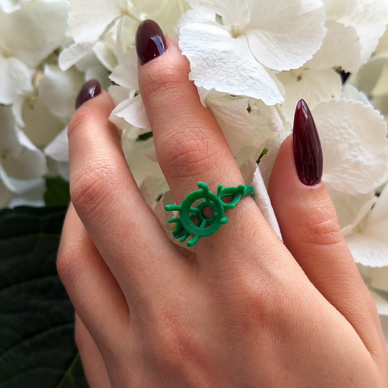Close-up of a hand with dark red nail polish, wearing a green ring resembling a ship's steering wheel, holding white hydrangea flowers.