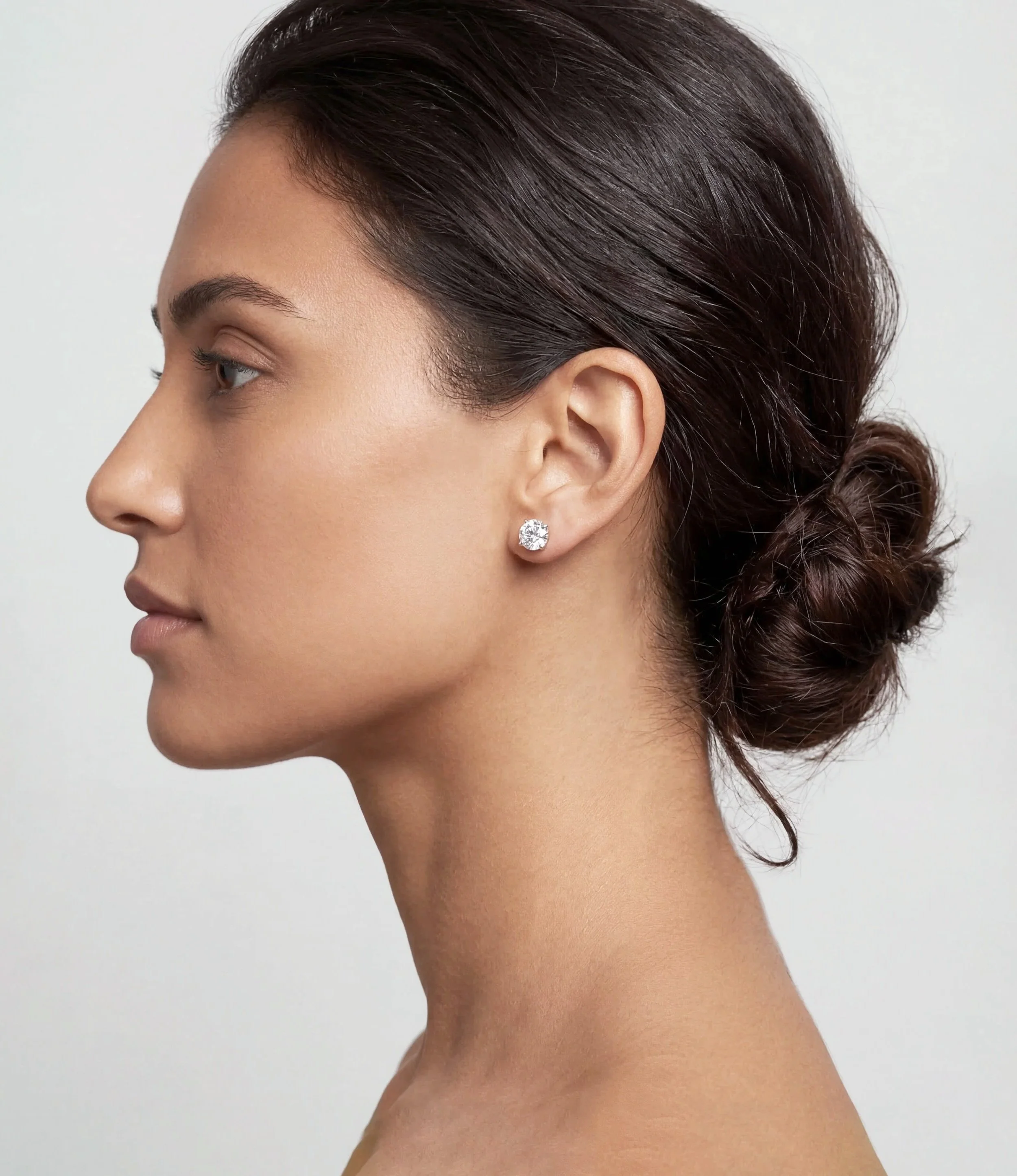 Side profile of a woman with dark hair styled in a low bun, wearing a diamond stud earring, with smooth skin and a neutral background.