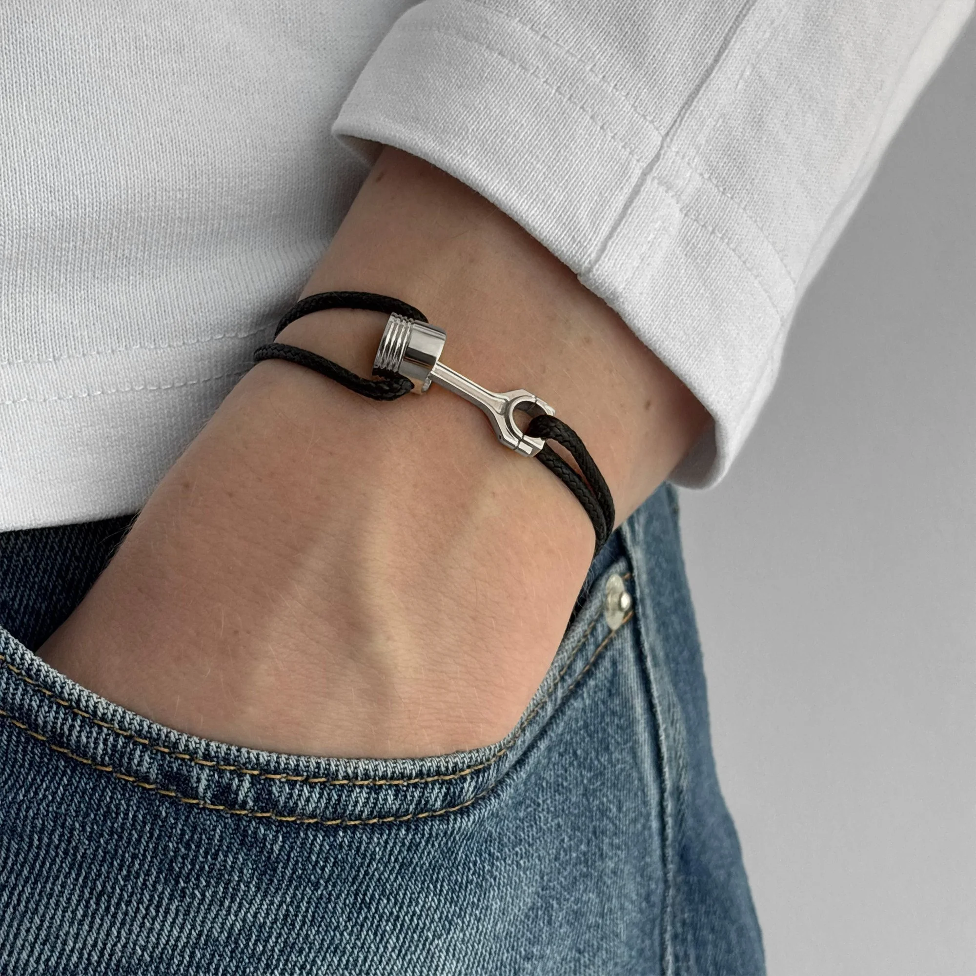 Person wearing a white long sleeve shirt and blue jeans, showing a black cord bracelet with a silver piston-shaped clasp on their wrist.